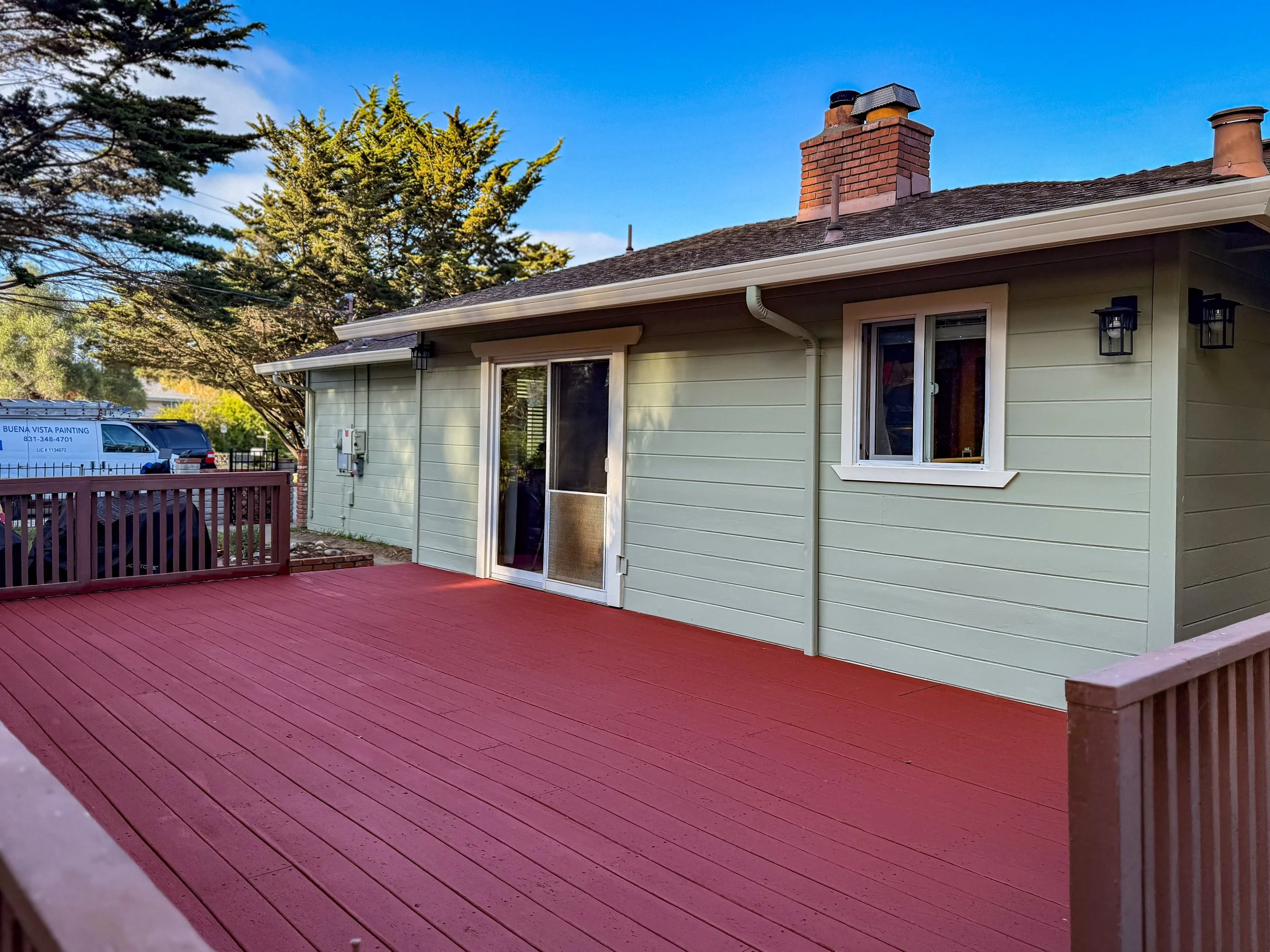 View of a backyard deck attached to a house with light green exterior siding, a sliding glass door, and a window. The deck has a reddish wood surface and a railing. There are trees and other houses visible in the background.