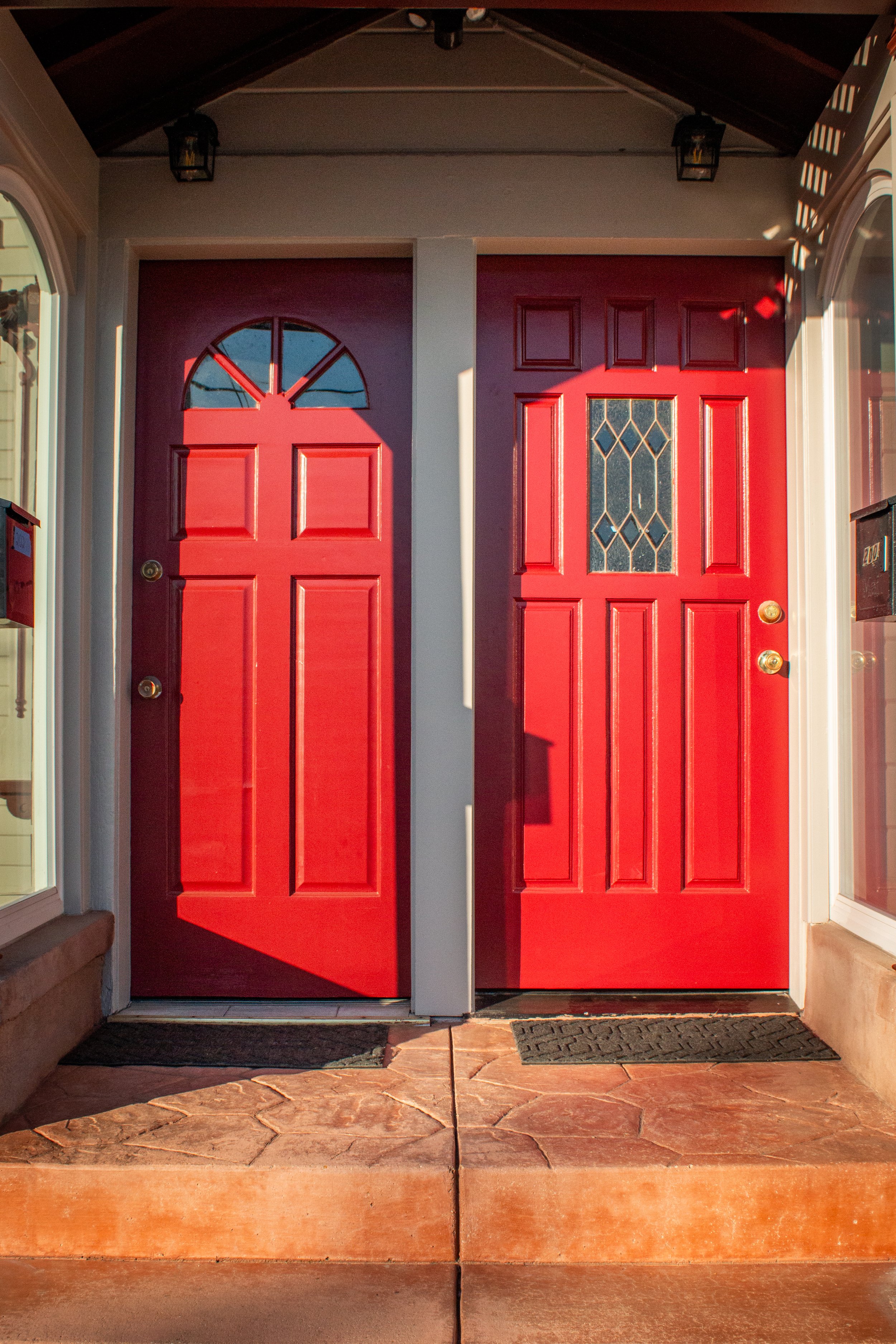 Two red front doors with glass window panels, situated side by side, with a small step and welcome mats in front.