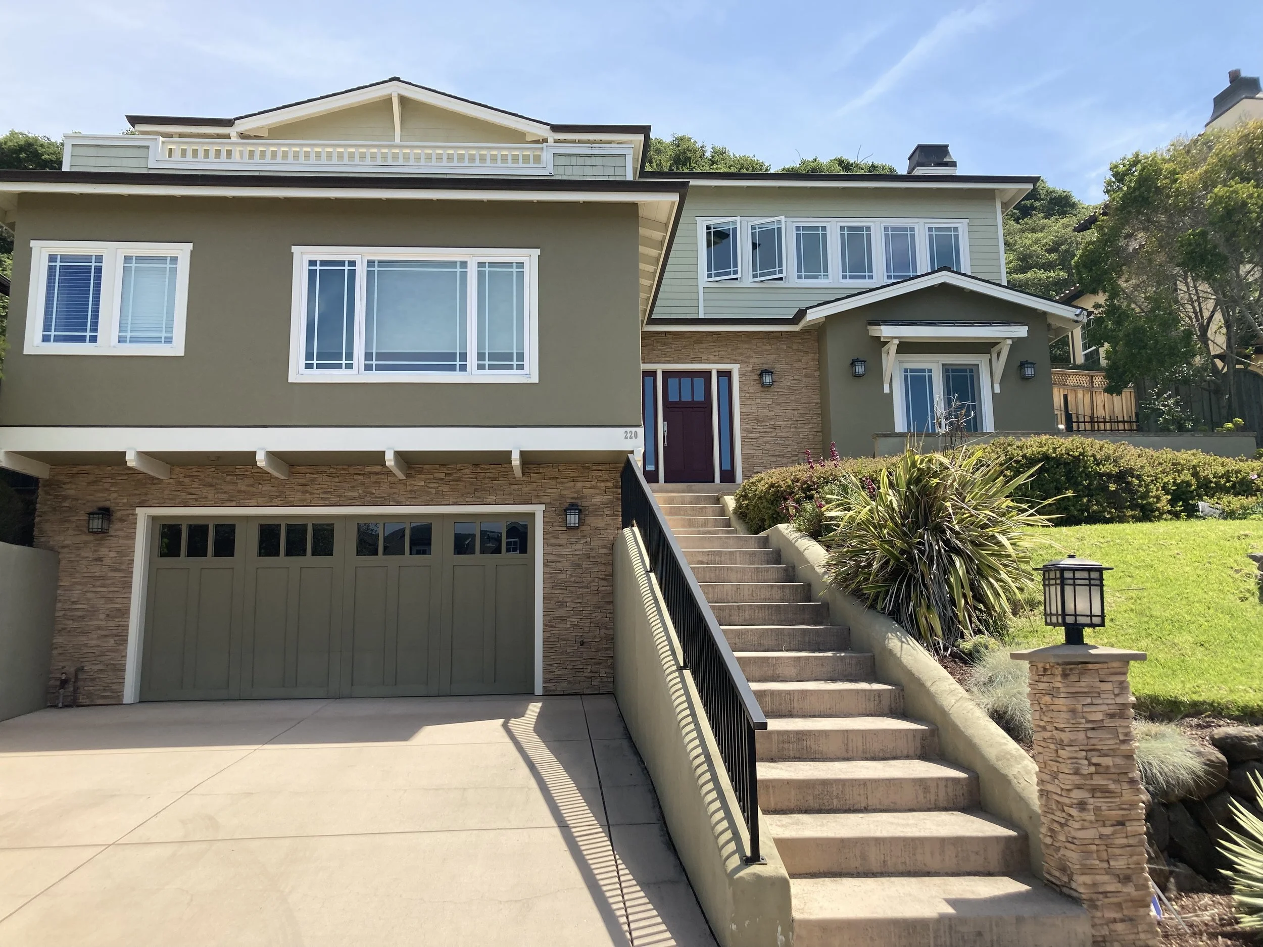Front view of a two-story house with stairs leading up to the front door, featuring a green garage door, multiple large windows, and landscaping with plants and bushes.