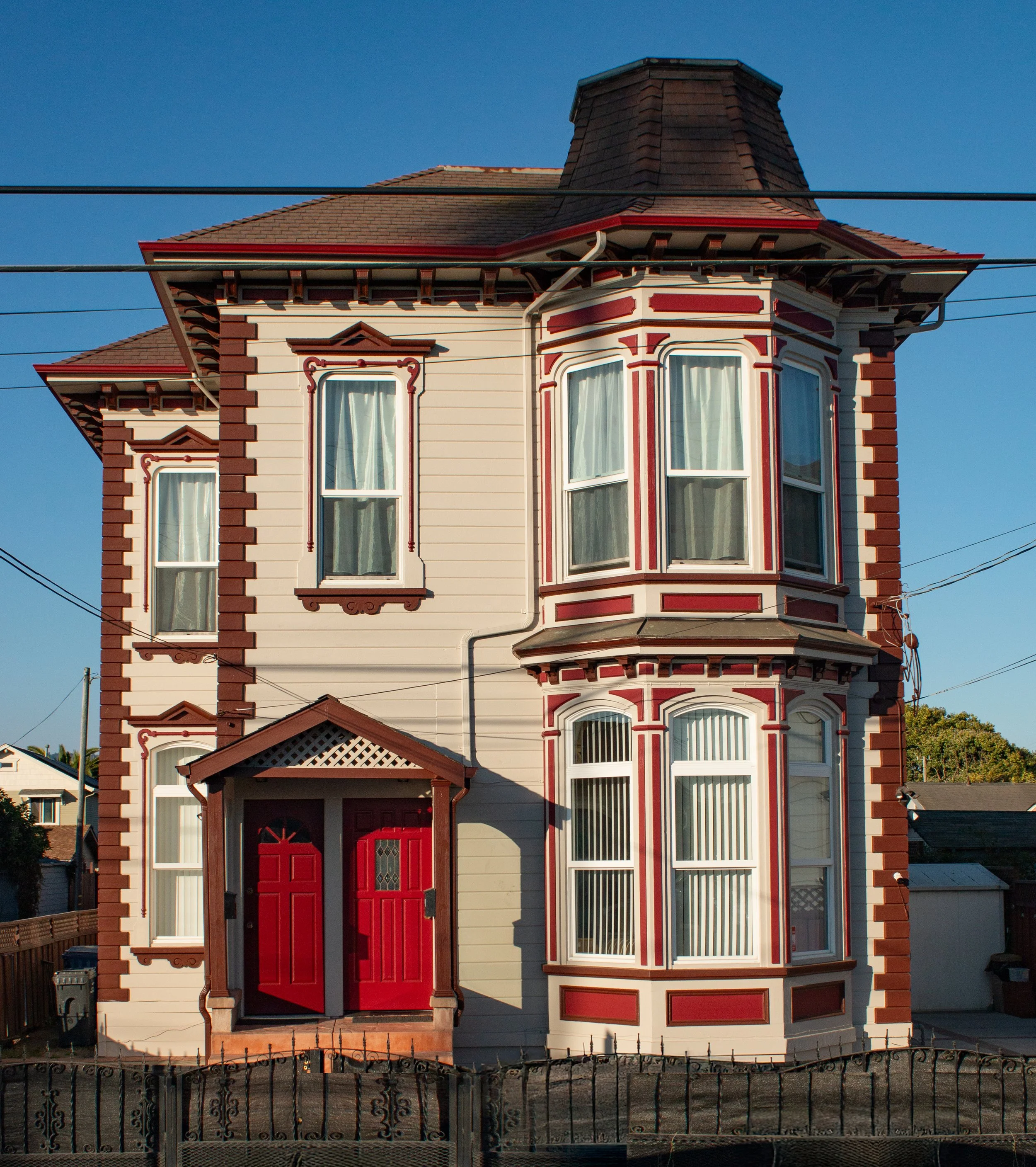 A three-story Victorian house with beige siding, red trim, and a gabled roof. The house features a prominent corner turret with large bay windows and a front porch with red double doors.