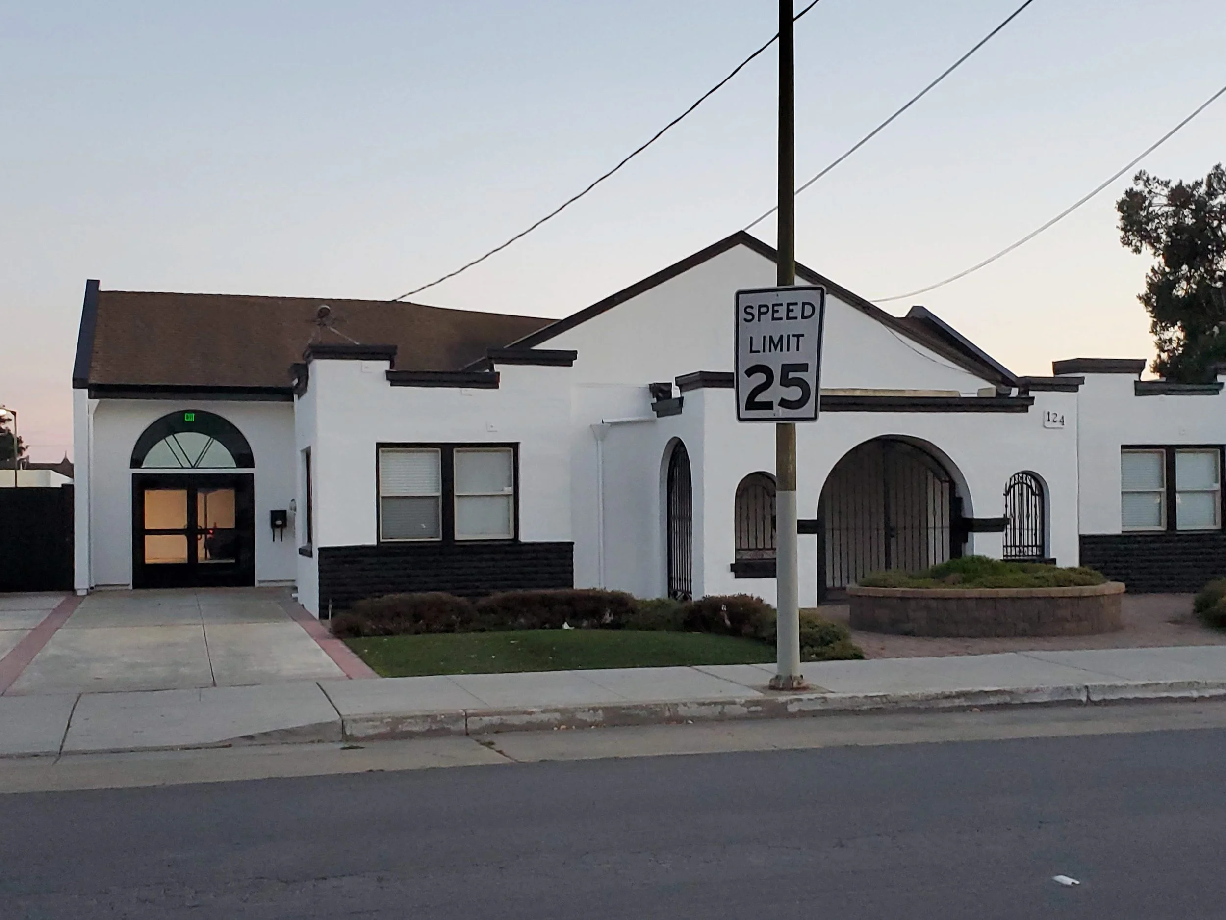 White building with black accents, arched windows, and a small front yard. Traffic sign indicating speed limit of 25 mph is in front of the building.