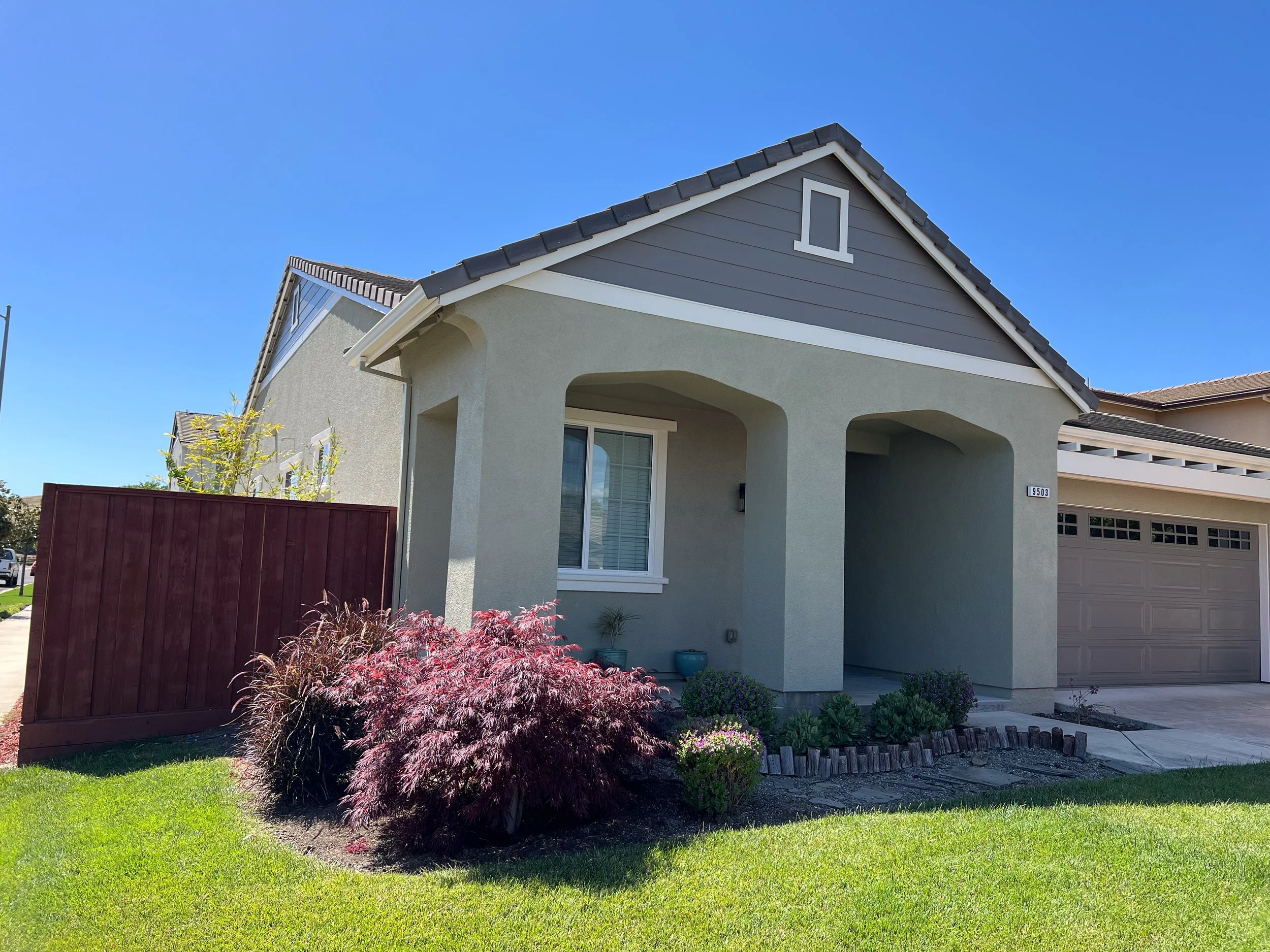 Front view of a modern house with a small front yard, landscaped with green grass, shrubs, and a red bush. The house has gray walls, white trim, and a gray garage door. A wooden fence is on the left side, and a blue sky is above.