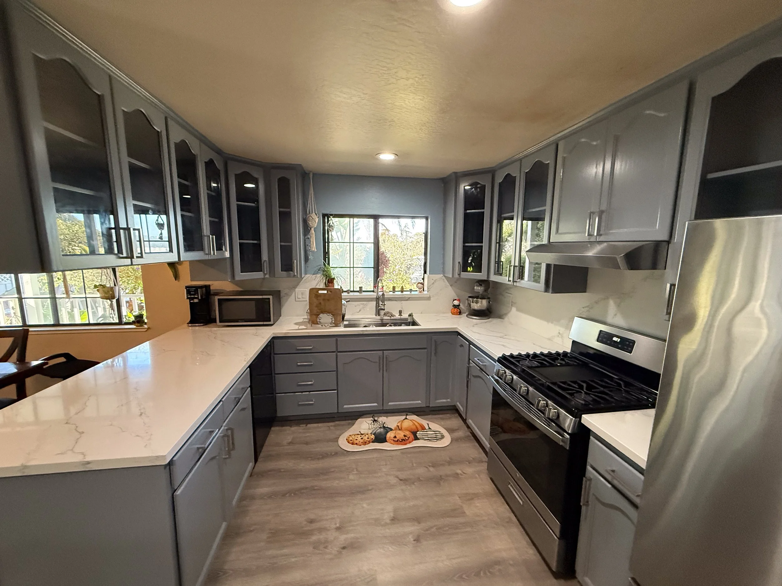 U-shaped kitchen with gray cabinets, white marble countertops, and stainless steel appliances, including a stove and refrigerator, with a window above the sink and a small decorative rug with pumpkins on the floor.