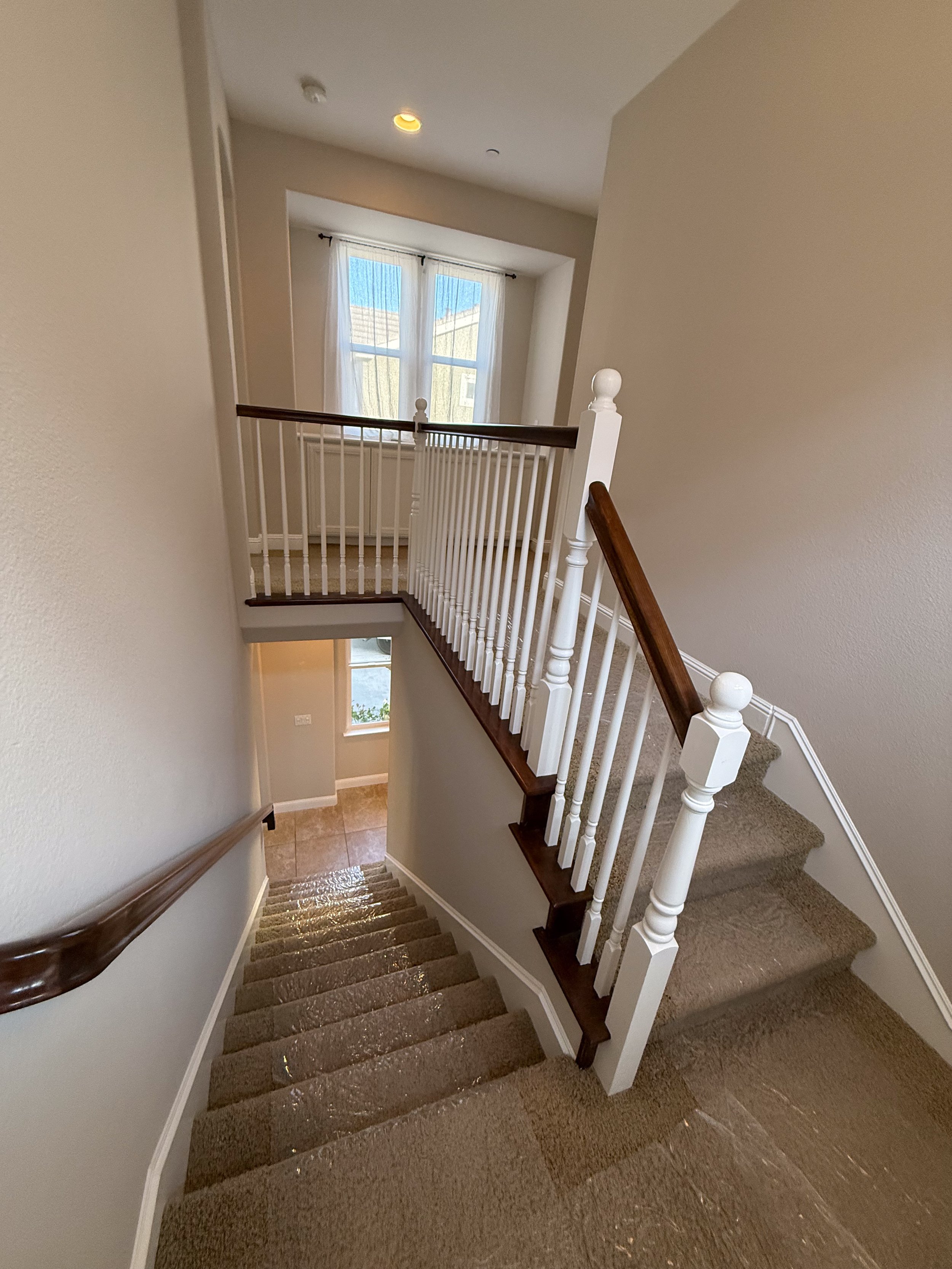 Interior view of a staircase with beige carpet, white walls, and a wooden handrail, leading down to a small landing with a window and a door.
