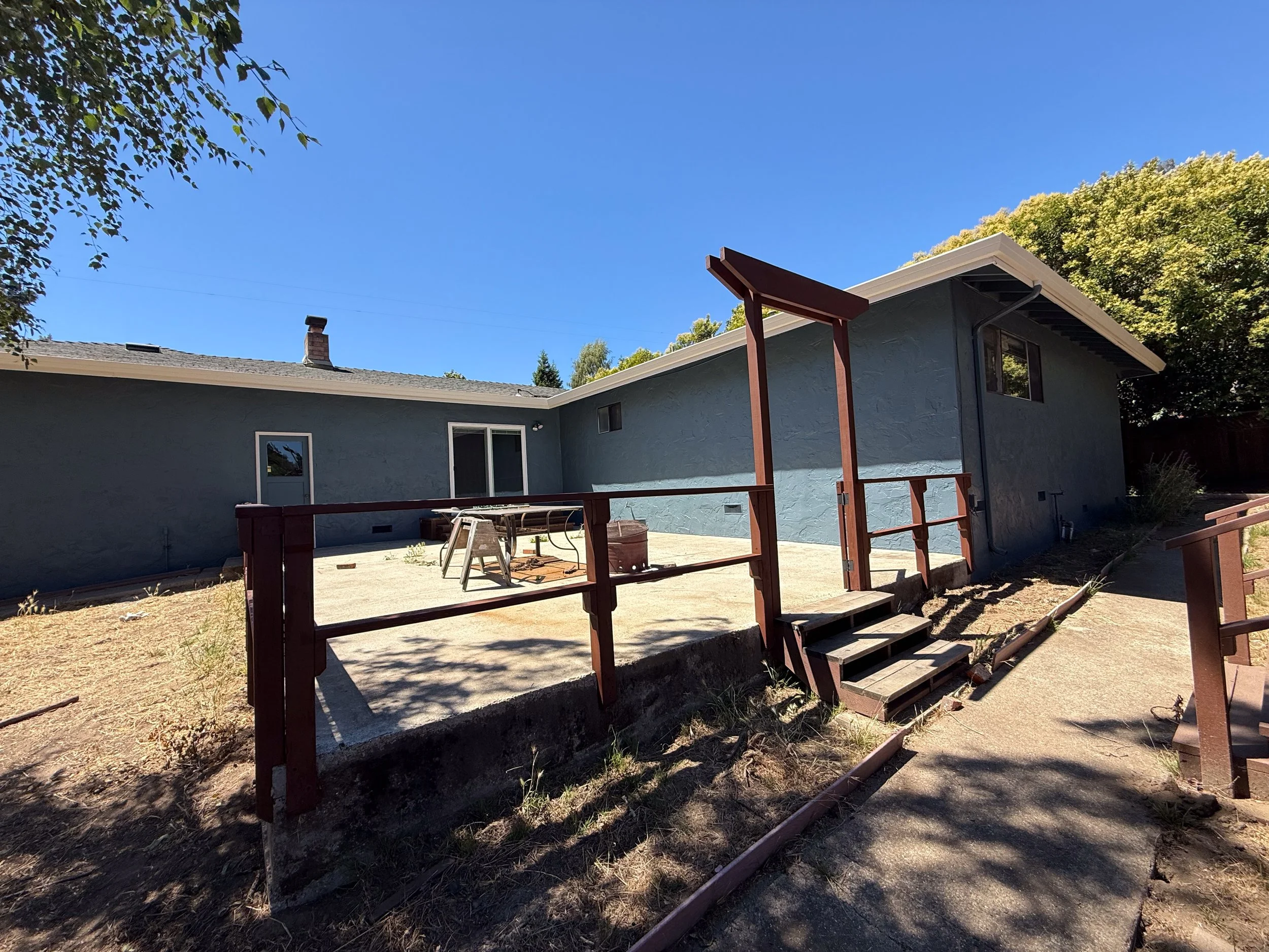 Backyard patio with a small set of wooden stairs, fence railing, two outdoor chairs, a table, and a shed against the house under a clear blue sky.