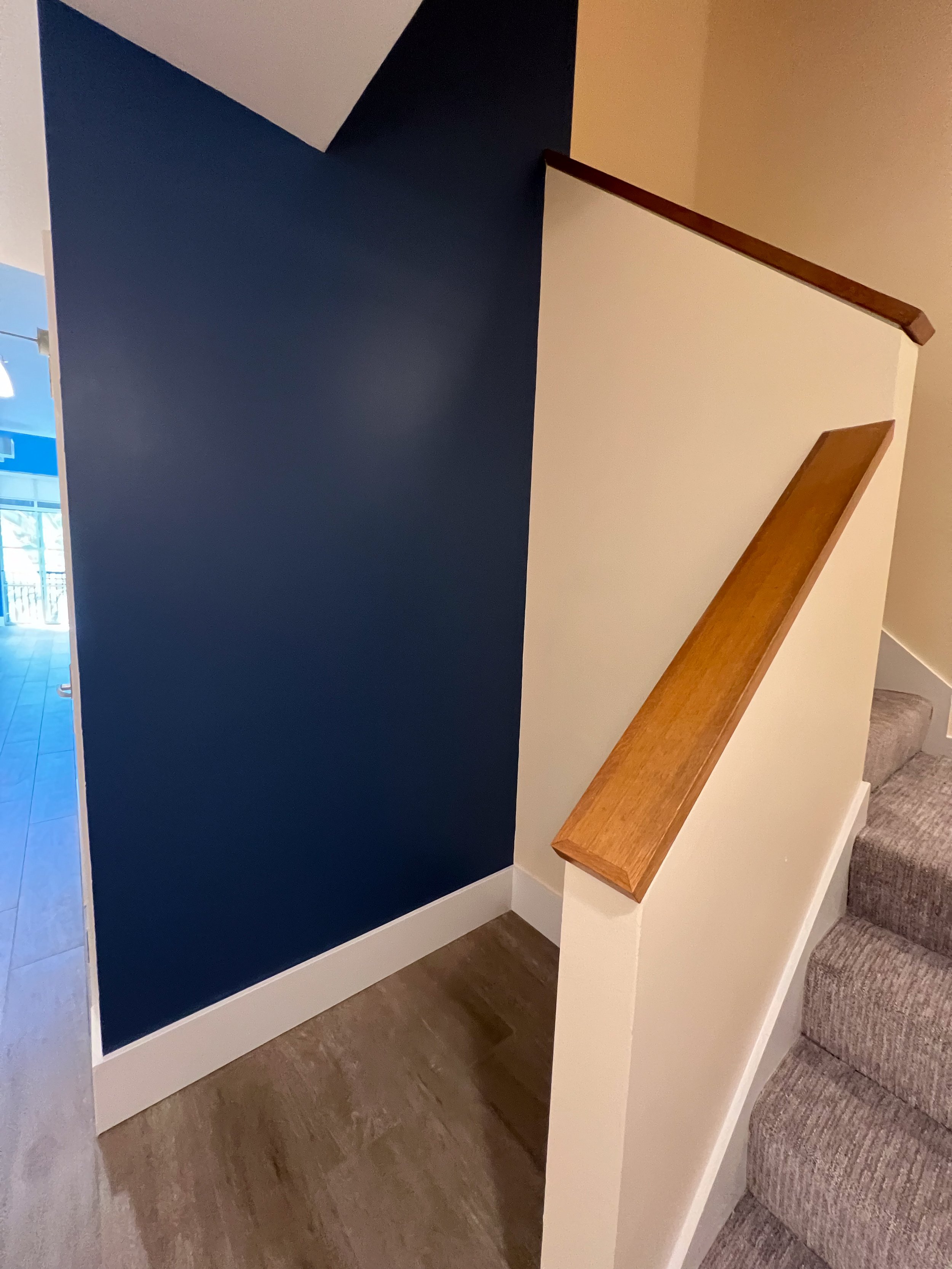Interior view of a staircase with carpeted steps, a beige wall with a wooden handrail, and a contrasting dark blue accent wall.
