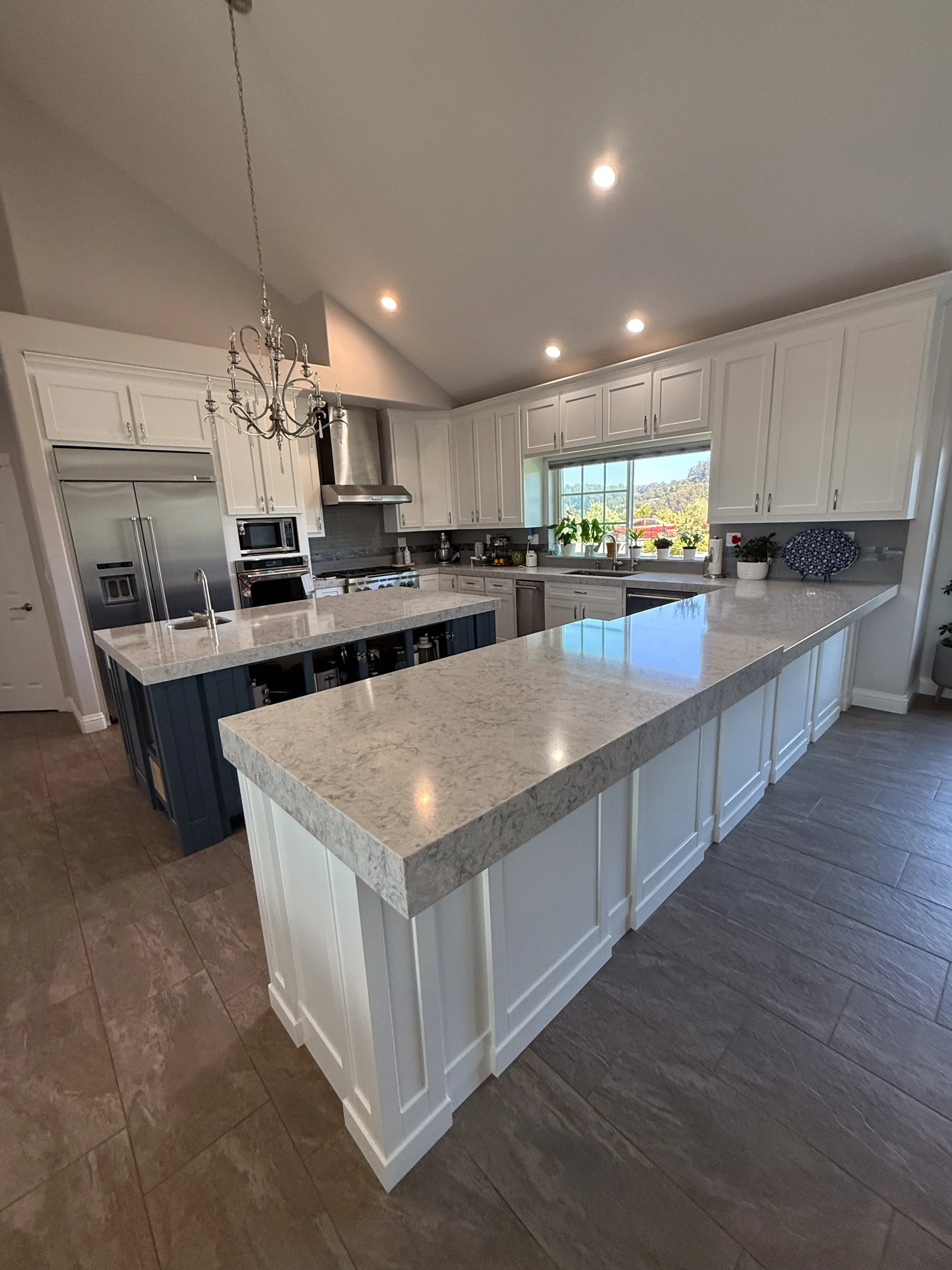 Modern kitchen with white cabinets, granite countertops, a central island, and stainless steel appliances. Large window with a view of trees, and a chandelier hanging above the island.
