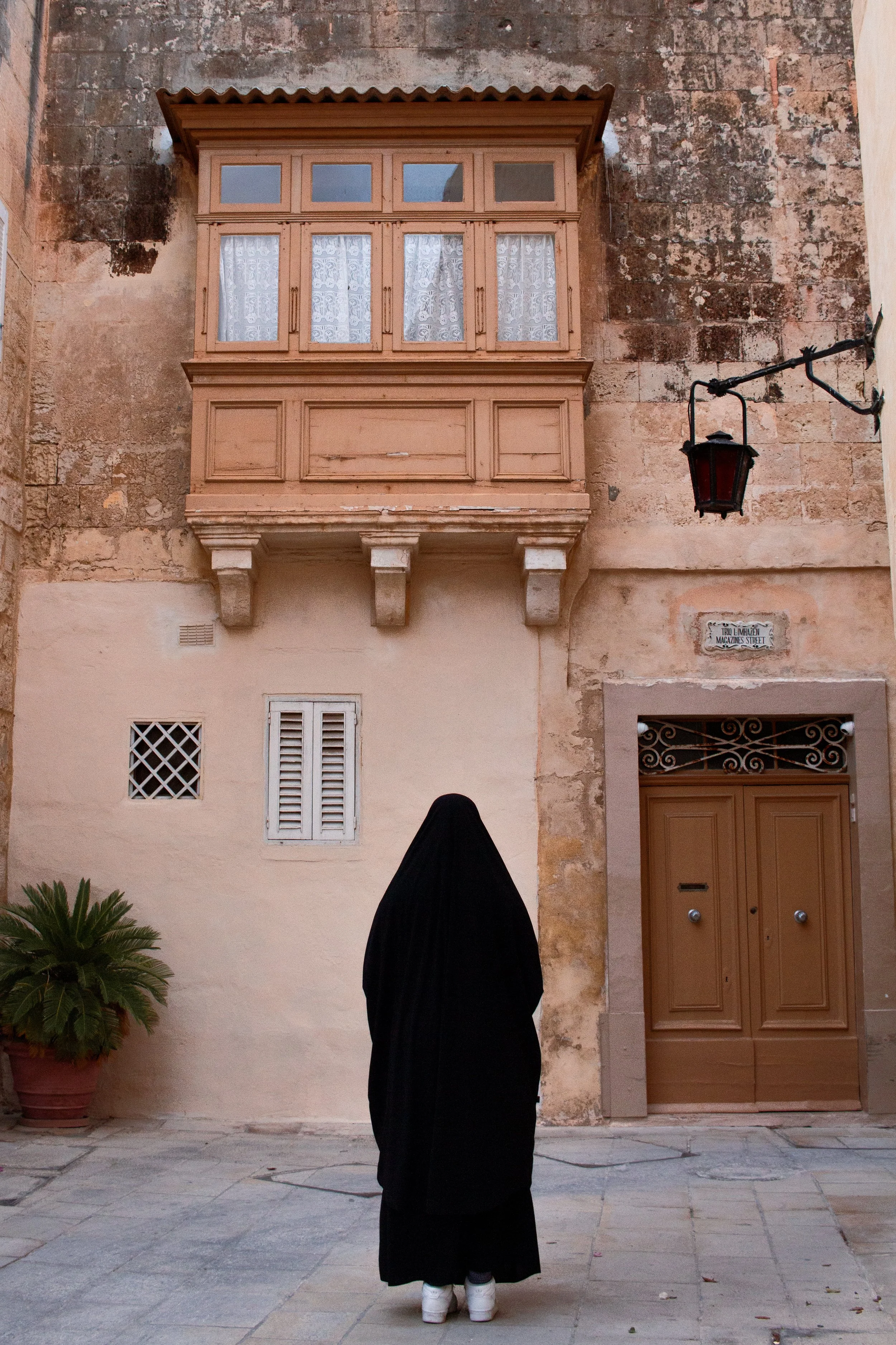 A woman dressed in black, standing with her back, in a narrow street with old buildings, potted plant, and a street sign that reads 'Trio Lumbaren, Magazine Street'.