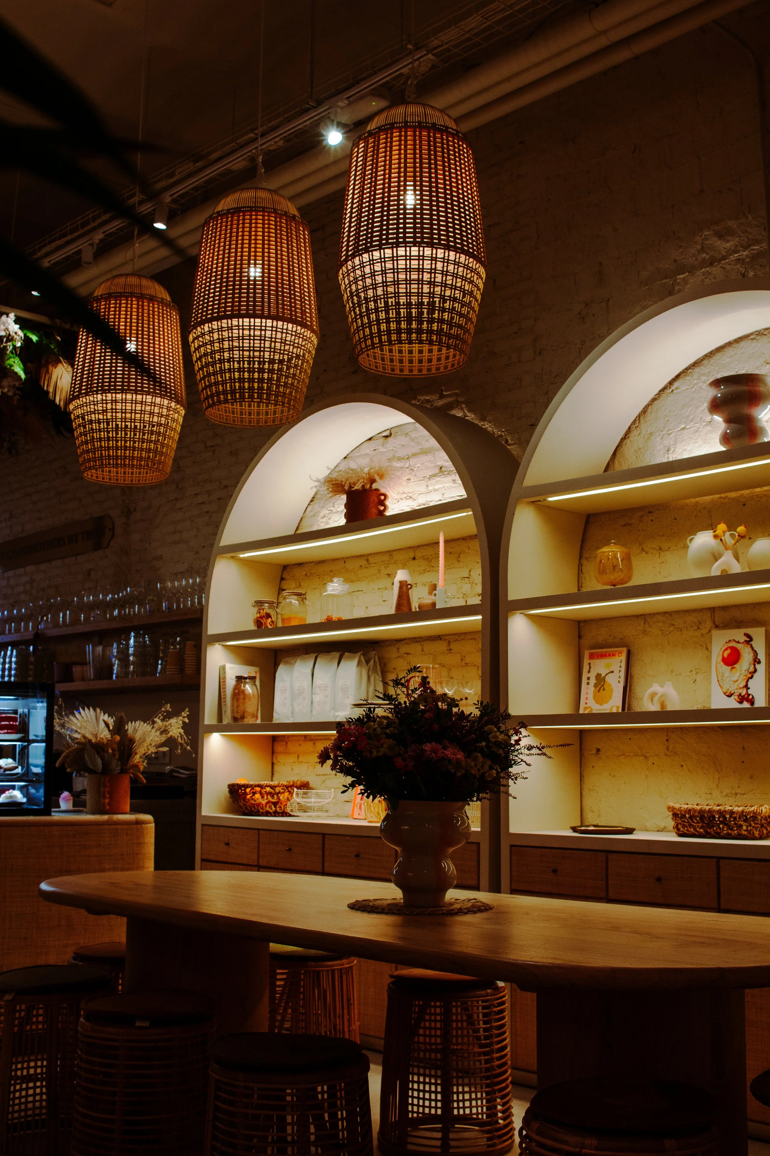 Interior of a cozy cafe with warm lighting, wicker pendant lights, floral centerpiece on a wooden table, and built-in shelves with decorative items.