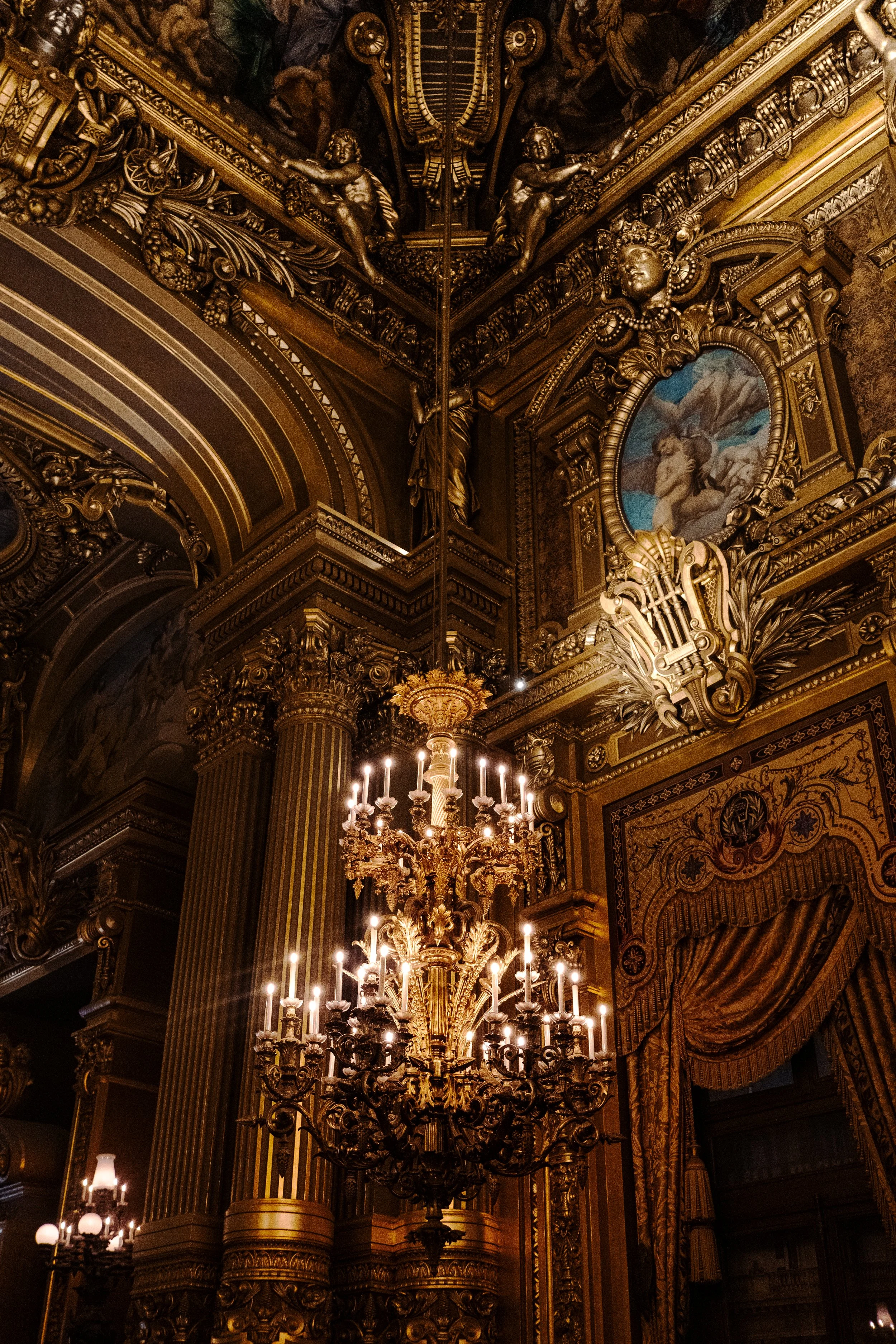 An ornate, gold-colored chandelier hangs from the ceiling of an opulent room with elaborate gold decoration, columns, and framed artwork.