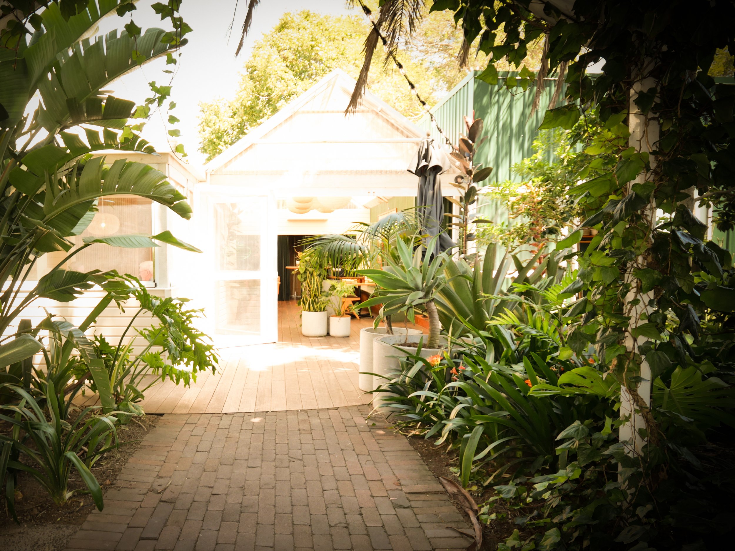 A restaurant  garden  made of bricks leading to a patio area with potted plants and an outdoor table, surrounded by lush green plants and trees, Lantern Papamoa.