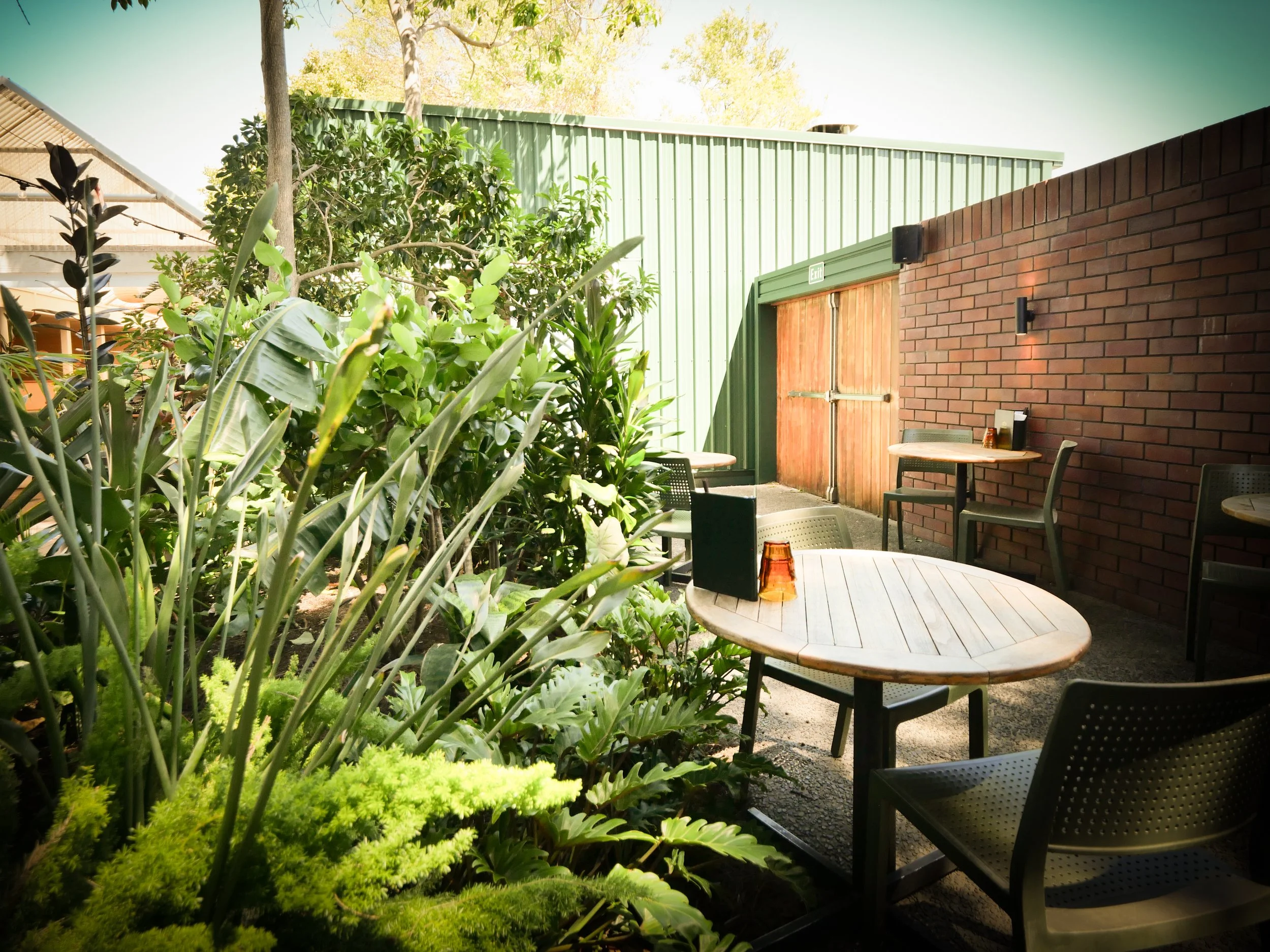 Outdoor patio area with round tables and chairs surrounded by green plants and trees, with a brick and metal building wall and door in the background.