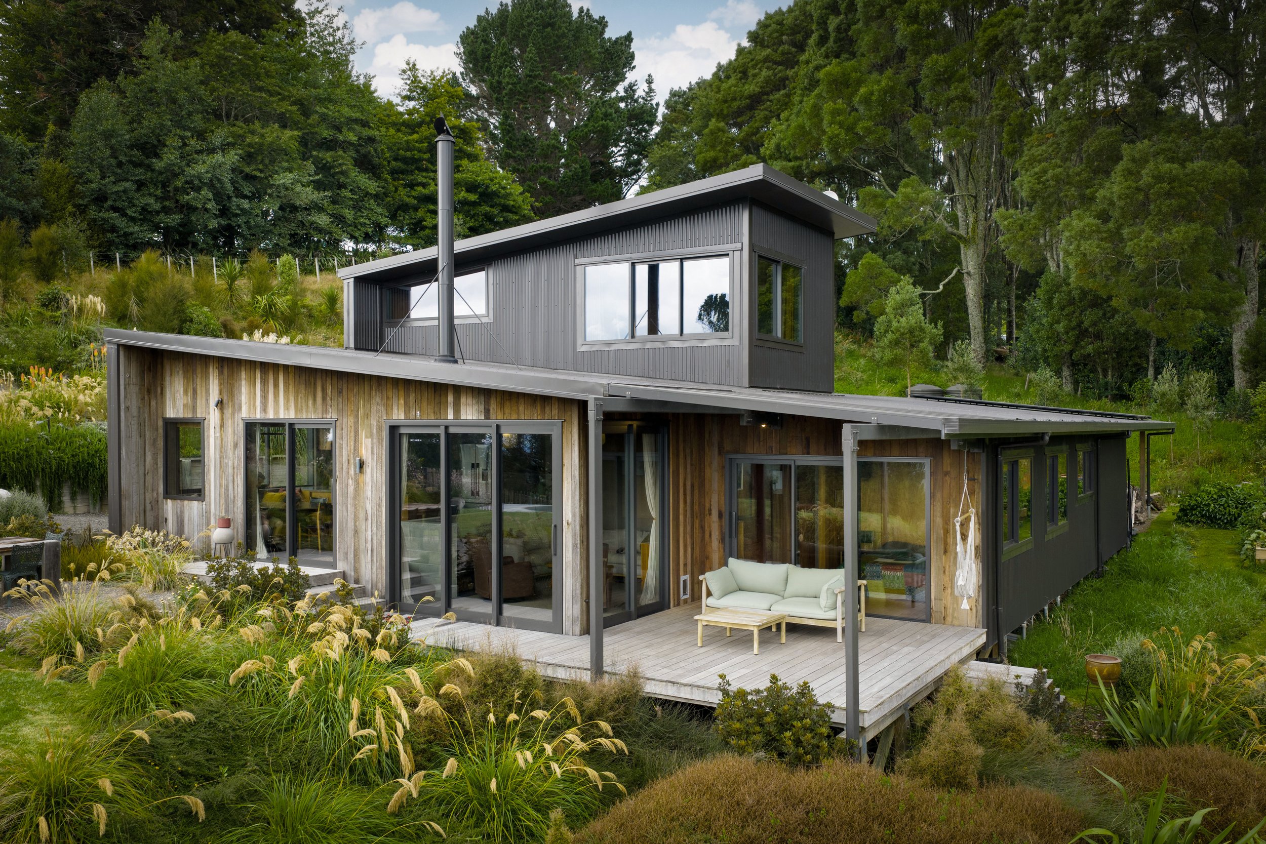 Modern two-story house, elevated and with large windows featuring recycled Australian hardwood cladding and decking and set in to a mostly NZ native planting scheme.