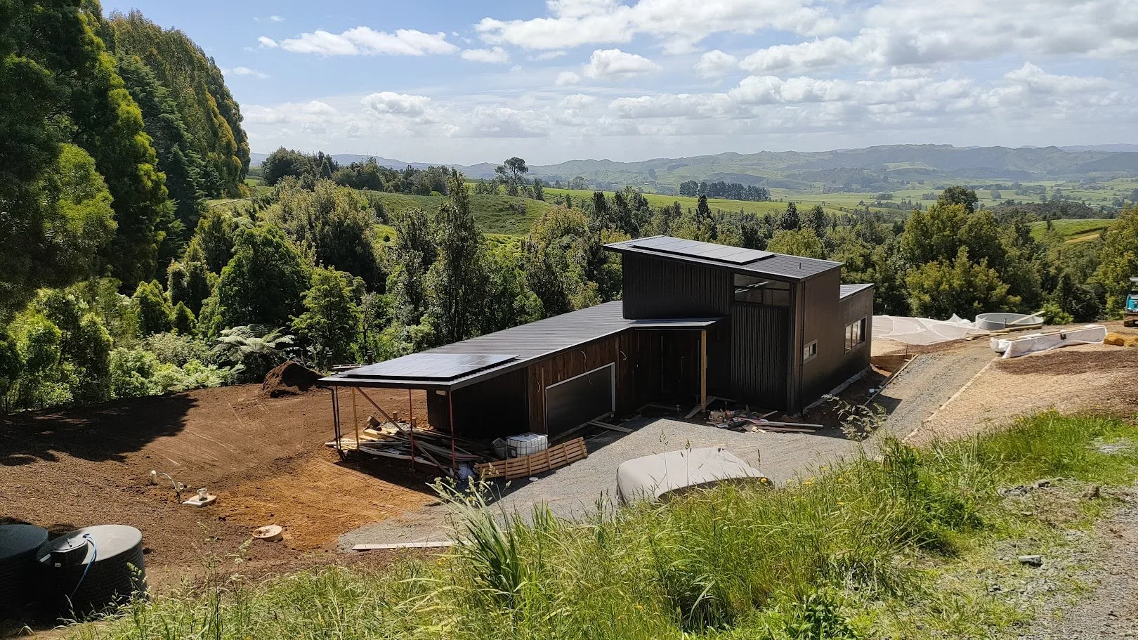 Under construction modern black house on a hillside with a scenic landscape of trees and rolling hills in the background under a partly cloudy sky.