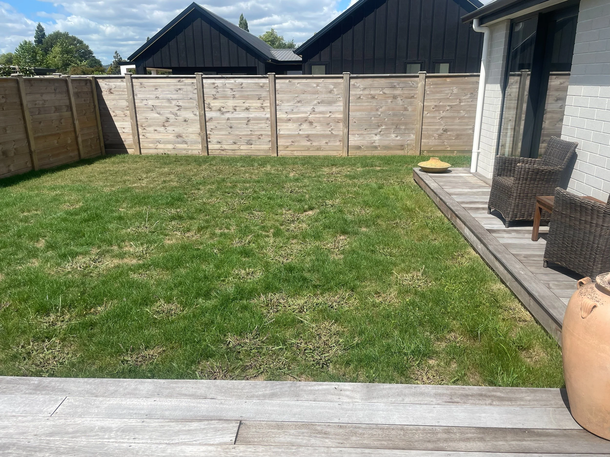 View of a small backyard with a grassy lawn, wooden deck, and a white brick house. A wooden fence surrounds the yard, and neighboring houses with dark roofs are visible in the background. Patio furniture and decorative items are on the deck.