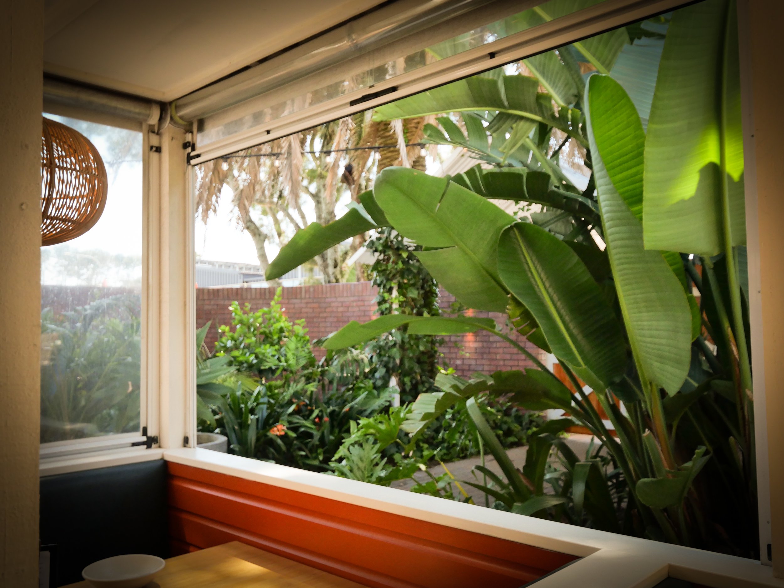 Open window with a view of a lush green garden and trees outside, with a wicker lampshade partially visible on the left.