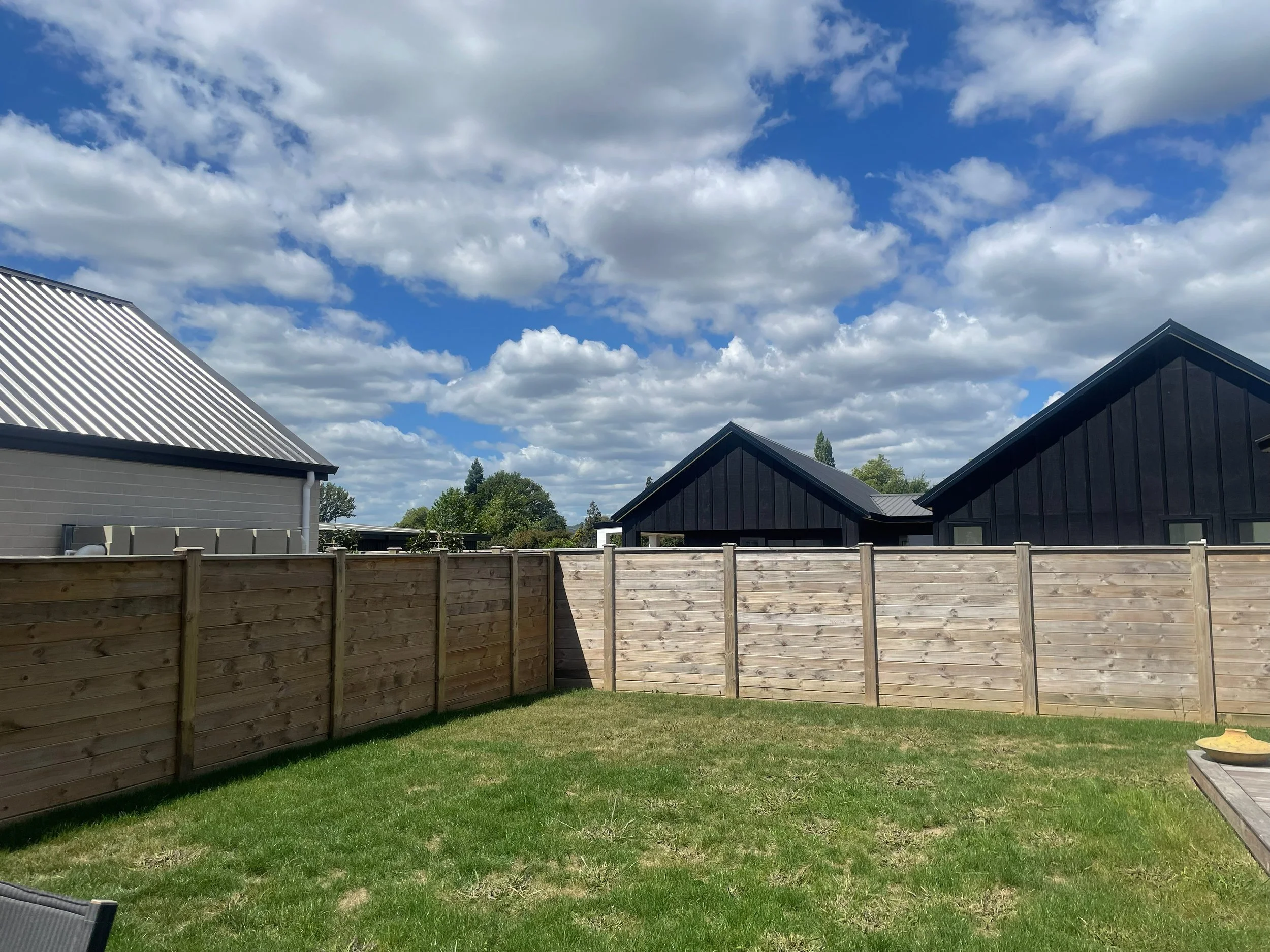 A backyard with a wooden privacy fence, a well-maintained grass lawn, and houses with sloped roofs in the background under a partly cloudy sky.