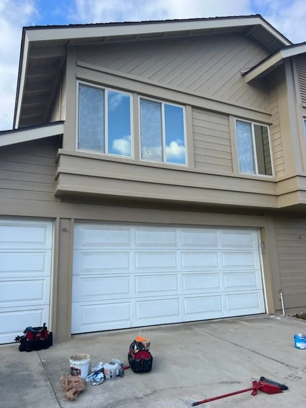 Front view of a two-story house with a white garage door, beige siding, and large windows reflecting the blue sky and clouds. There are some tools and items on the driveway.