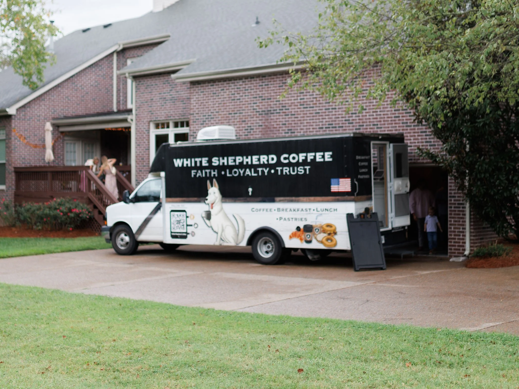 A White Shepherd coffee food truck parked in a driveway in front of a brick house with a porch. The truck advertises coffee, breakfast, lunch, pastries, and features a picture of a white Shepherd dog with a QR code, an American flag, and images of do