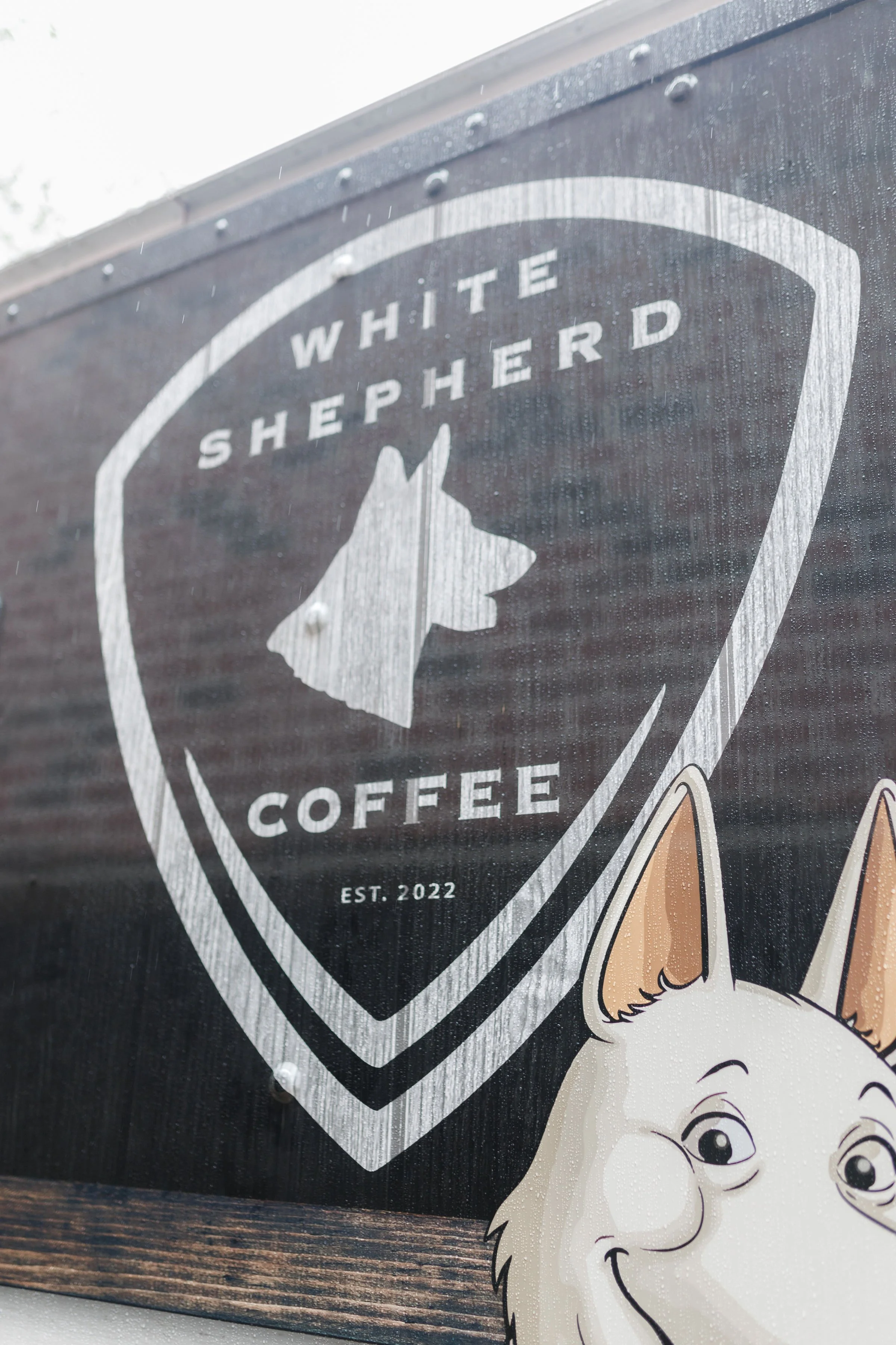 Close-up of a sign for White Shepherd Coffee, established in 2022, featuring a shield logo with a profile of a white shepherd dog and part of a cartoon dog illustration at the bottom right corner.