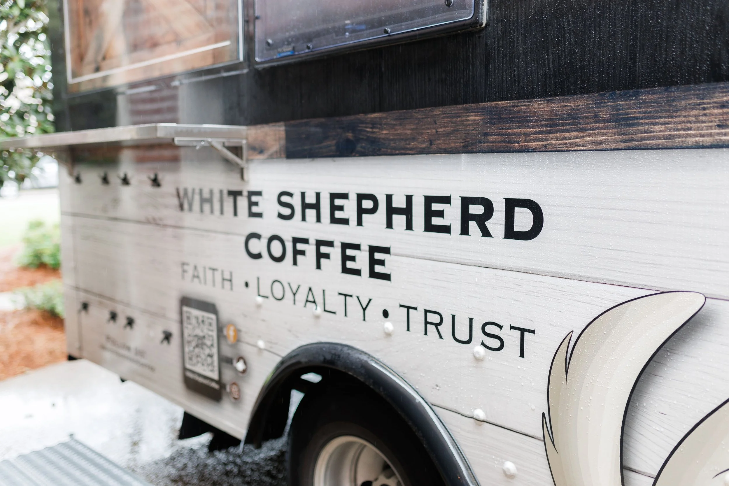 A coffee food truck with the name 'White Shepherd Coffee' and words 'Faith, Loyalty, Trust' on the side, featuring a white and brown exterior with a feather illustration.