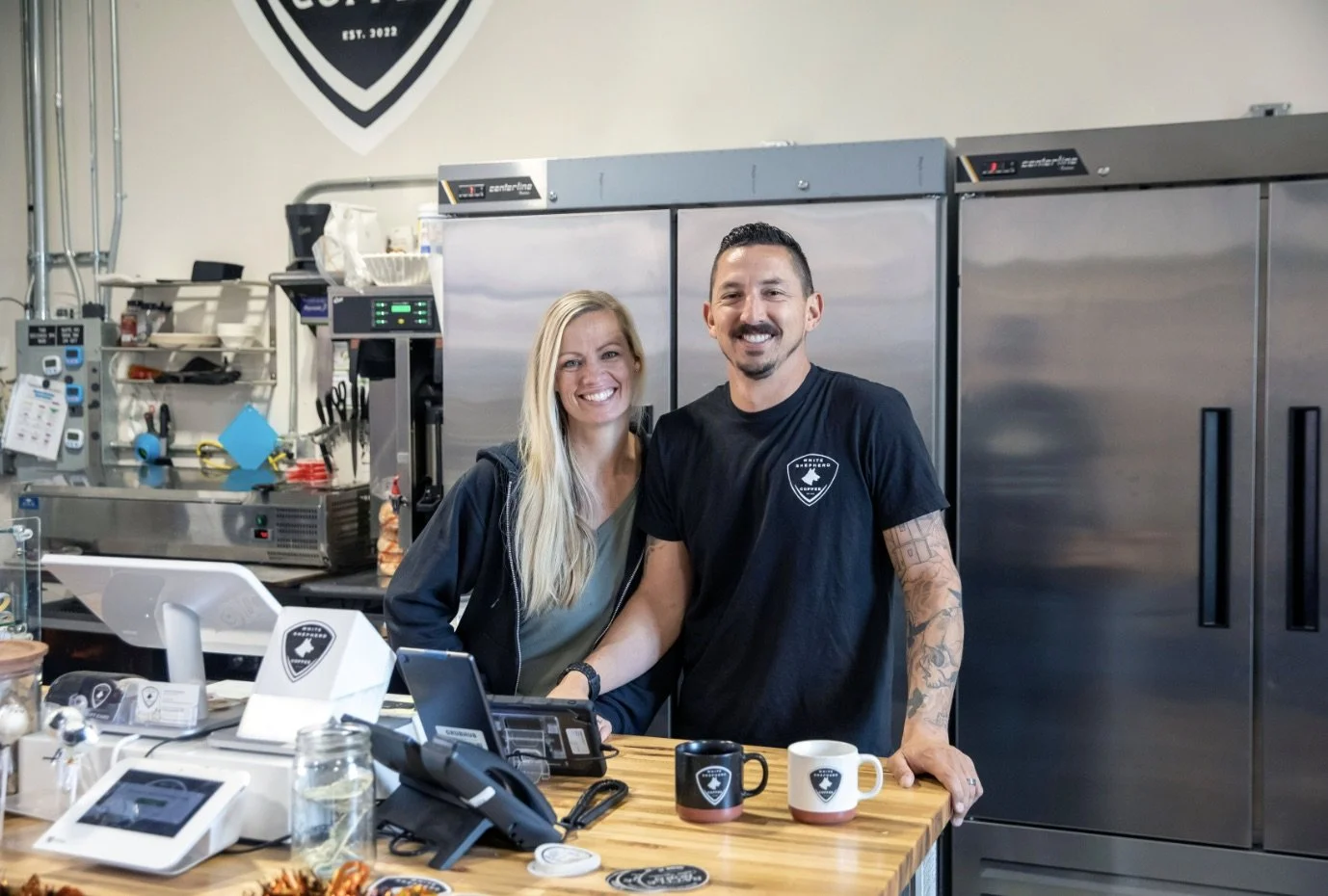 Two smiling people, a woman and a man, standing behind a wooden counter in a commercial kitchen or café, with coffee mugs on the counter and various equipment around them.
