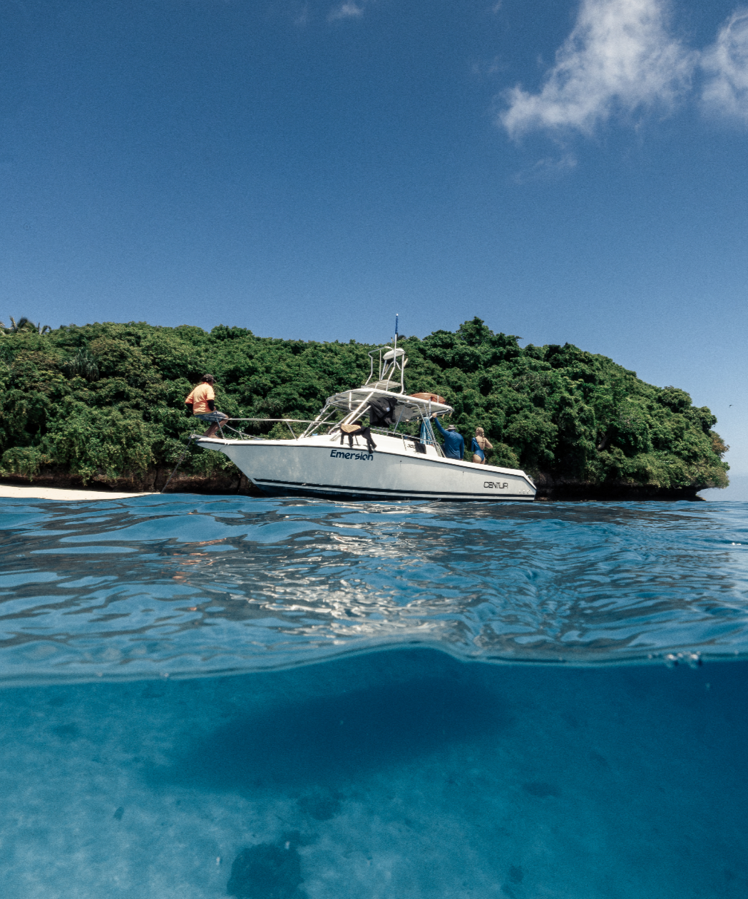 A boat named 'Emersion' anchored near a lush, green island with a bright blue sky overhead and clear blue water below.