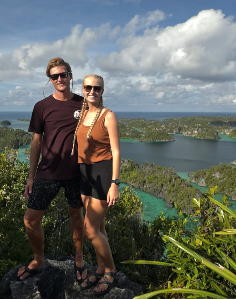 Andrew and Olivia are standing on a scenic ledge overlooking a lush, green, blue lagoon in Indonesia.