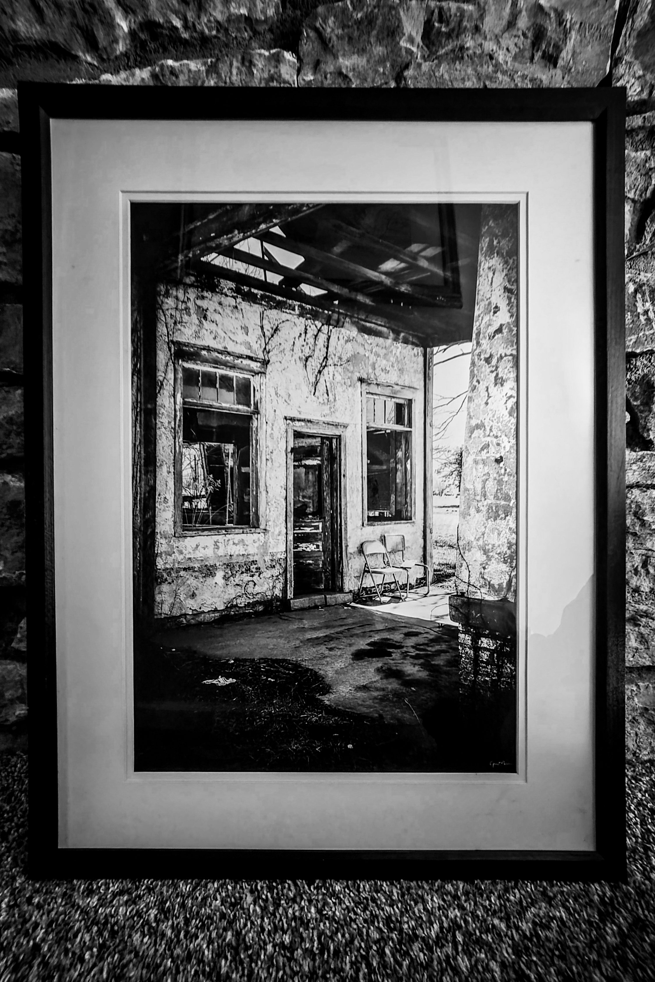 Black and white framed photograph of an abandoned building with broken windows, a chair, and outdoor trees.
