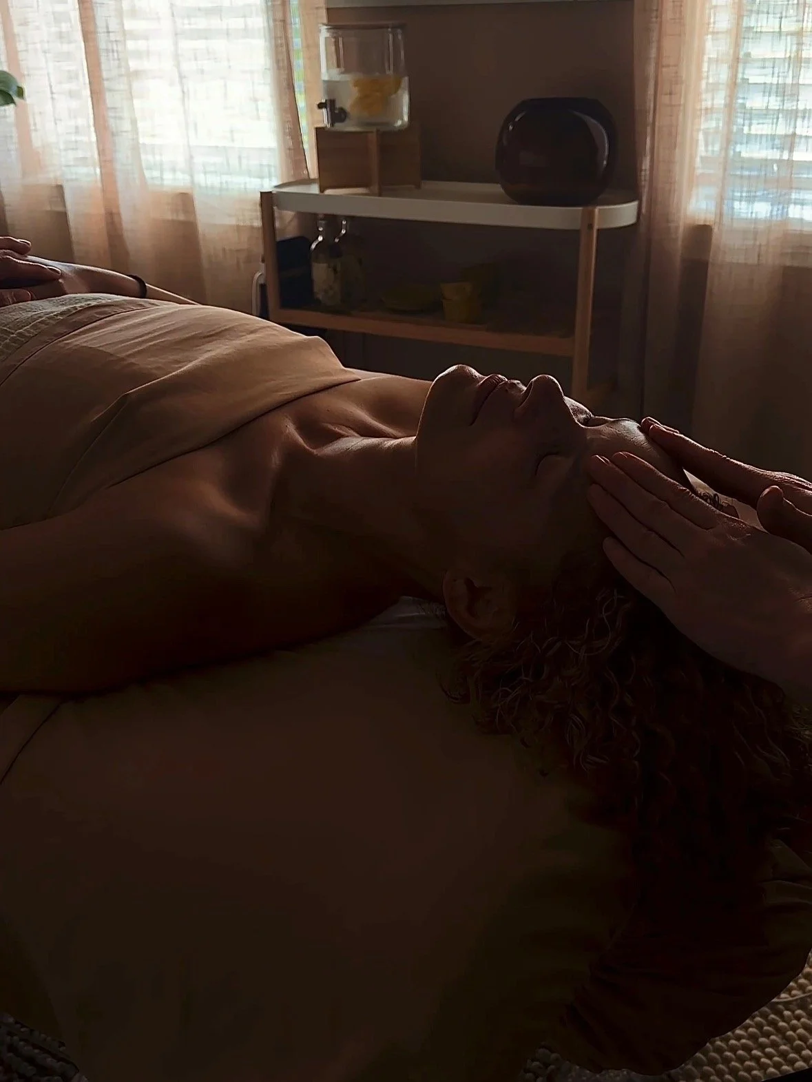 Person lying on their back in a dimly lit room, receiving a facial massage or treatment.