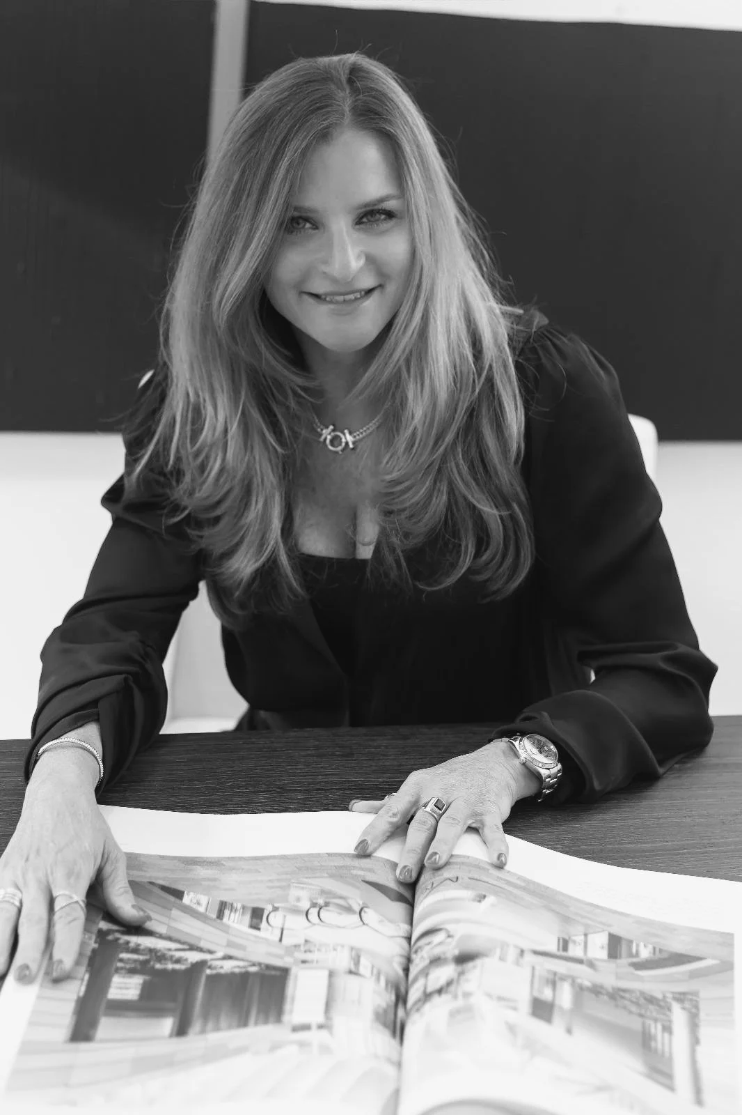 Black and white photo of a woman with long hair smiling with a magazine open on a table in front of her.