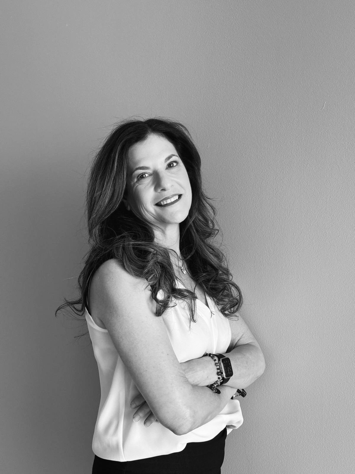 Black and white portrait of a woman with long wavy hair, smiling slightly, standing against a plain wall with arms crossed.