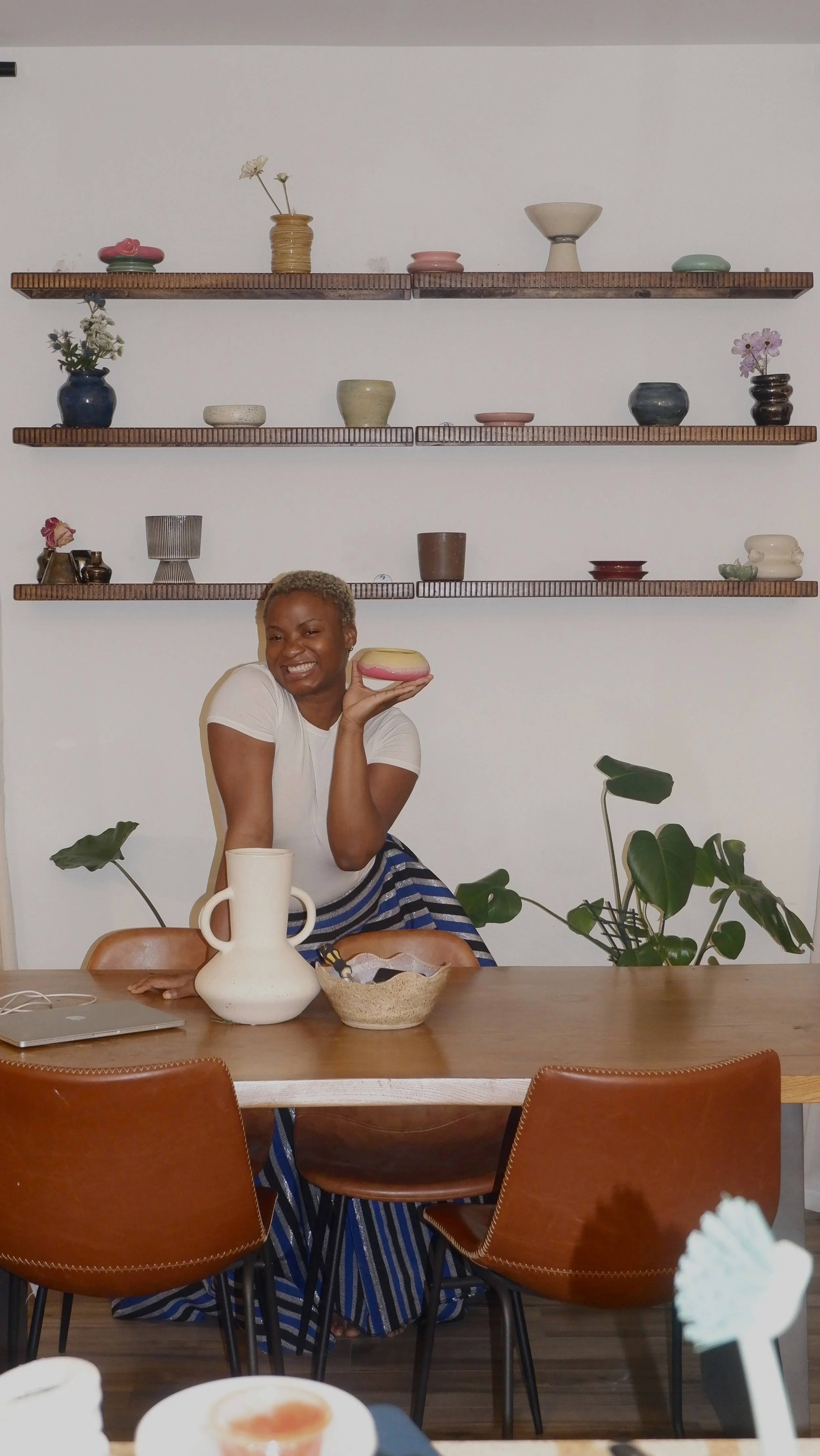 A woman with short blonde hair smiling and holding a ceramic bowl. She is seated at a wooden dining table with chairs, and behind her are three floating shelves with various decorative ceramics and vases, and green plants.