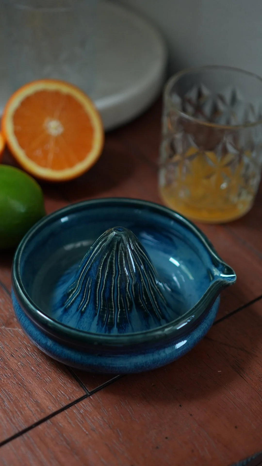 A blue ceramic juicer on a wooden surface, with half an orange, a lime, a glass with lemon slices, and a metal container in the background.