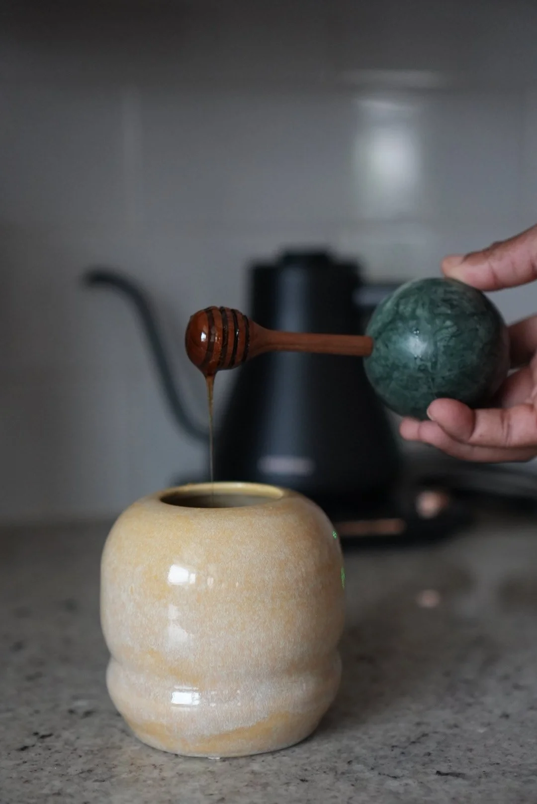 A hand holding a green stone ball and a wooden honey dipper, with honey dripping into a beige ceramic cup placed on a countertop, and a black kettle in the blurred background.