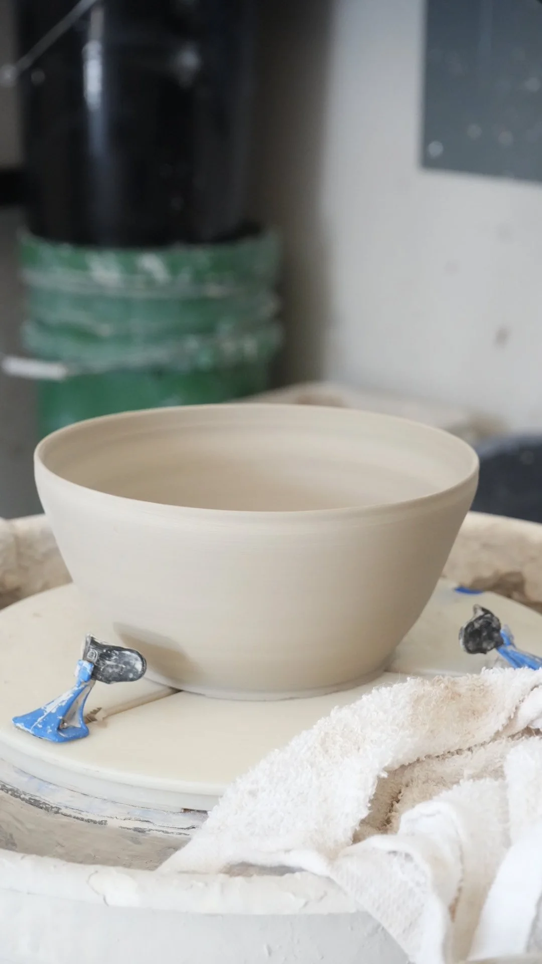 Unfinished clay bowl on a pottery wheel in a studio, with sculpting tools and a towel nearby.