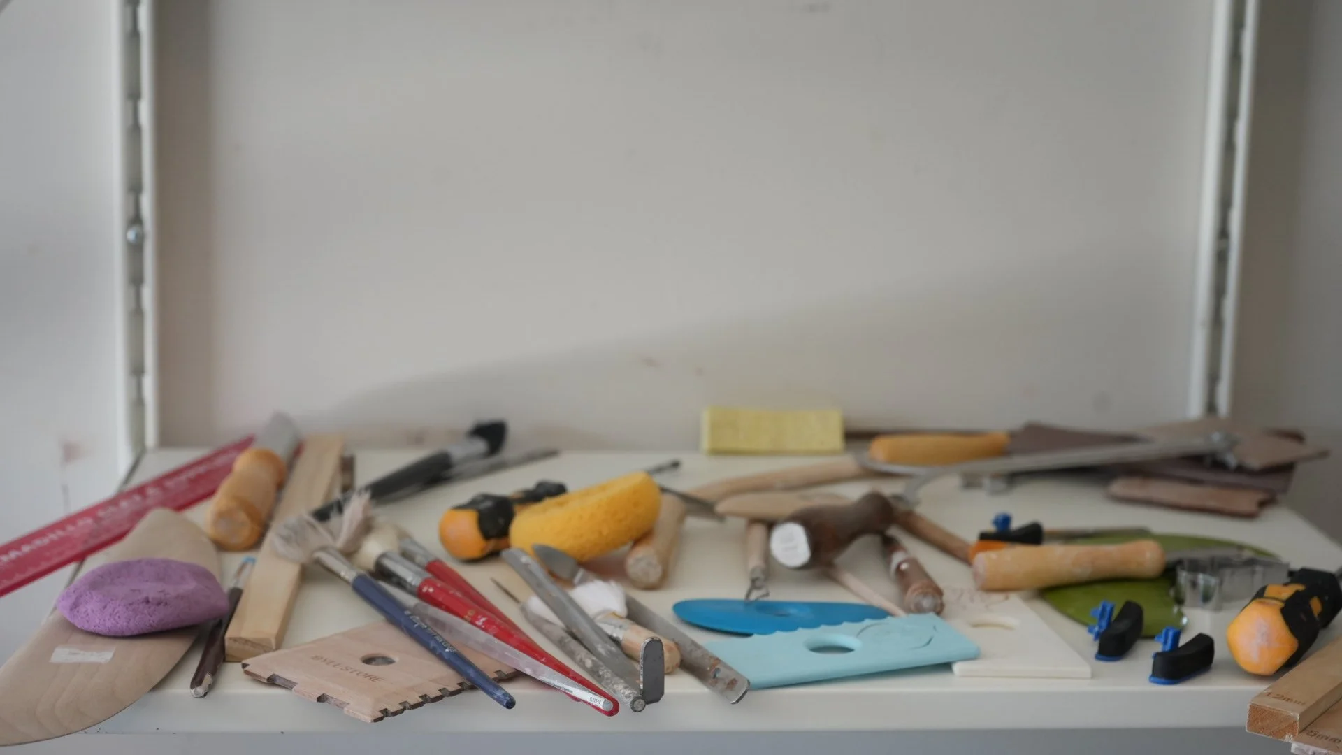 Tools and materials for woodworking and painting on a white surface, including paintbrushes, clamps, sanding sponge, ruler, and wood pieces.