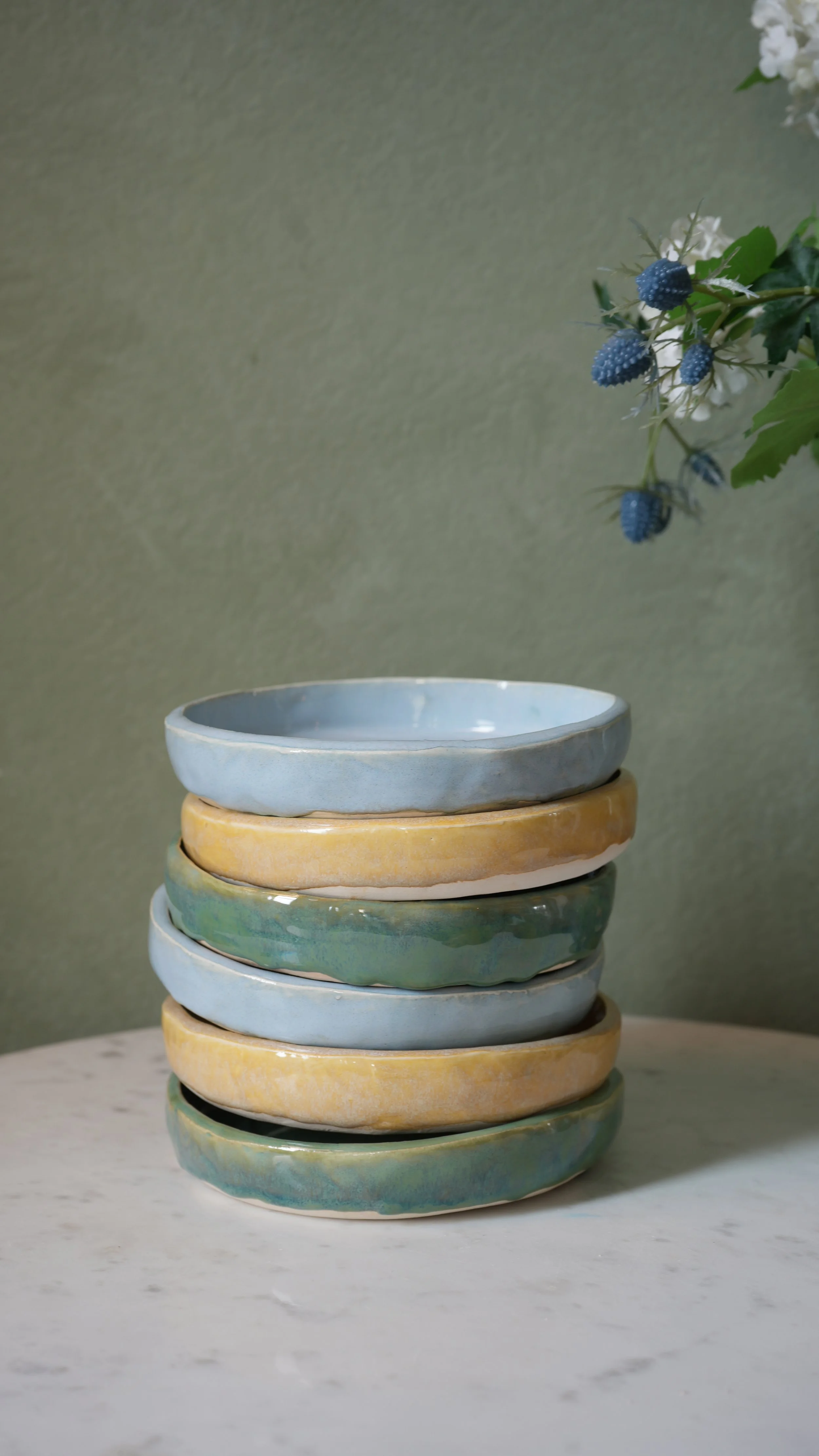 Stack of seven ceramic bowls in pastel blue, yellow, and green colors on a white marble surface with a green textured wall and a sprig of white and blue flowers in the background.