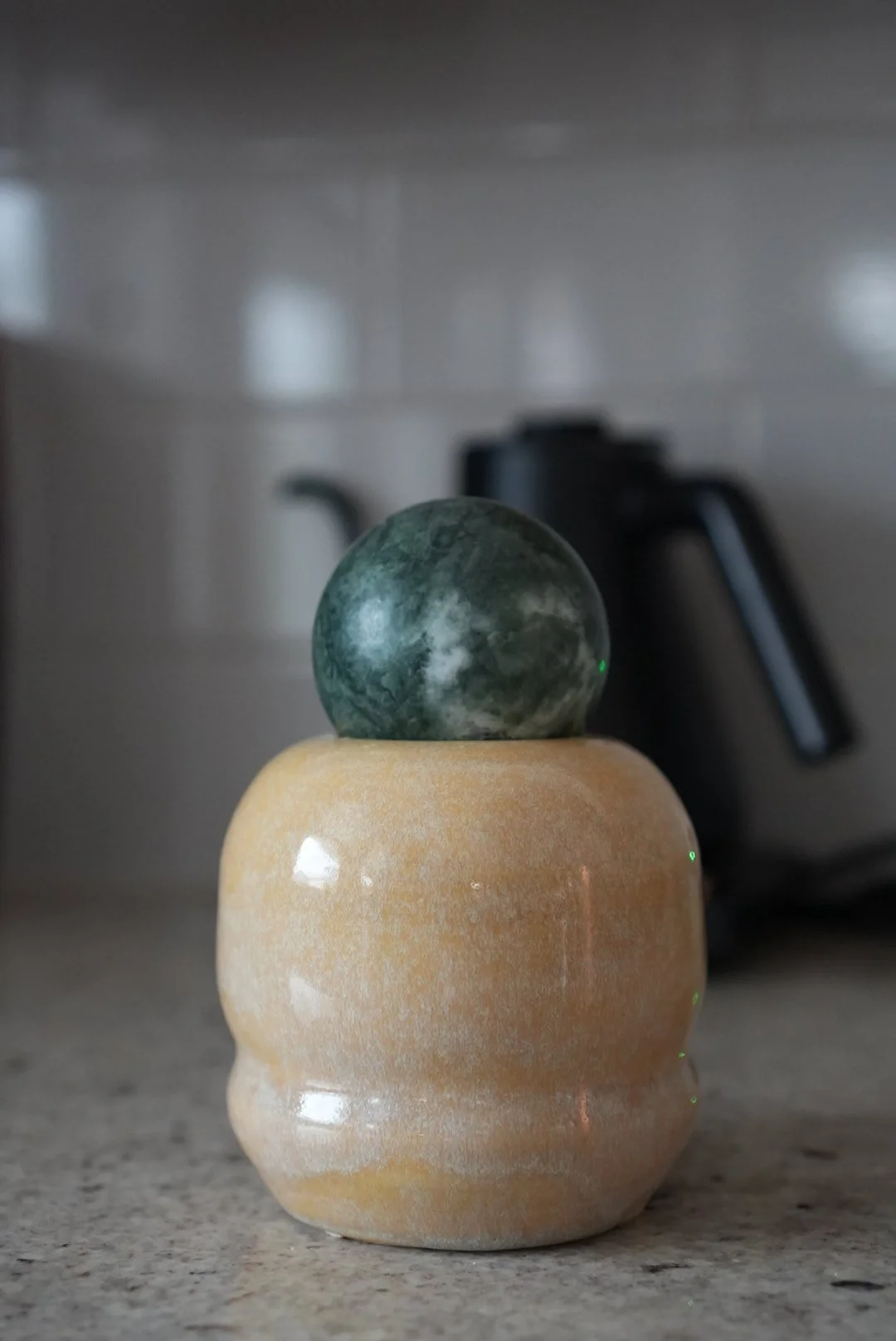 A beige ceramic vase with a rounded, ridged shape resting on a countertop. A dark green marble sphere is balanced on top of the vase. In the background, there is a black electric kettle on a tiled kitchen wall.