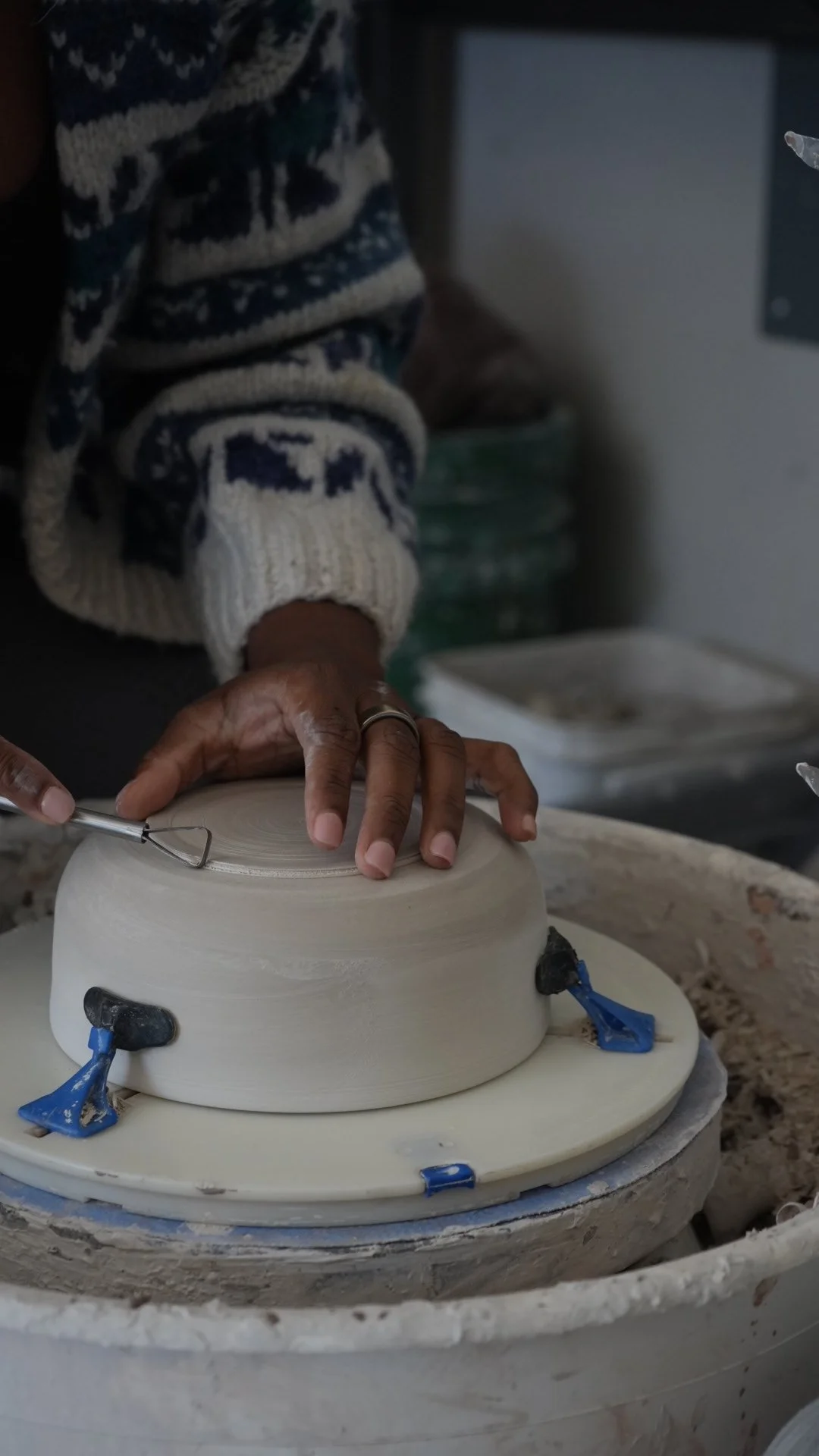 Person shaping a ceramic bowl on a pottery wheel, wearing a patterned sweater, with tools and clay surrounding.