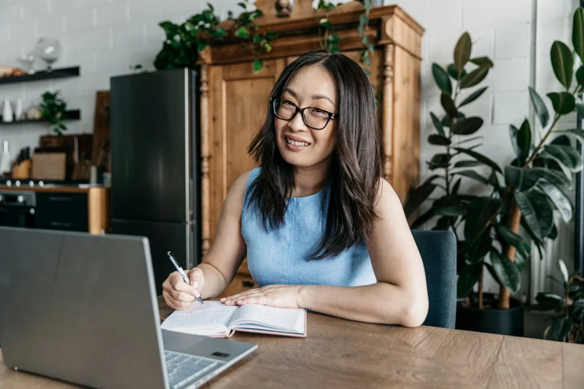 A woman sitting at a wooden table with a laptop and open notebook smiling.