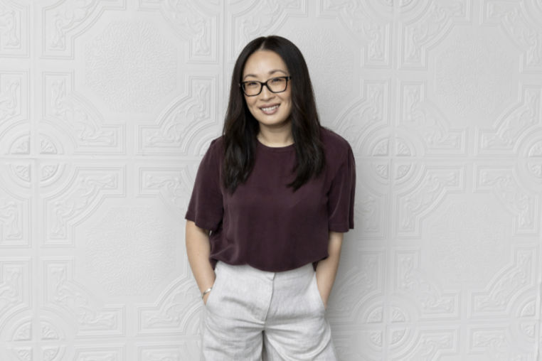 A woman with a purple top standing against a textured white wall.