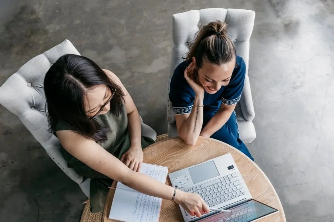 Two women sitting at a round wooden table, looking at a laptop, smiling and engaging with each other.