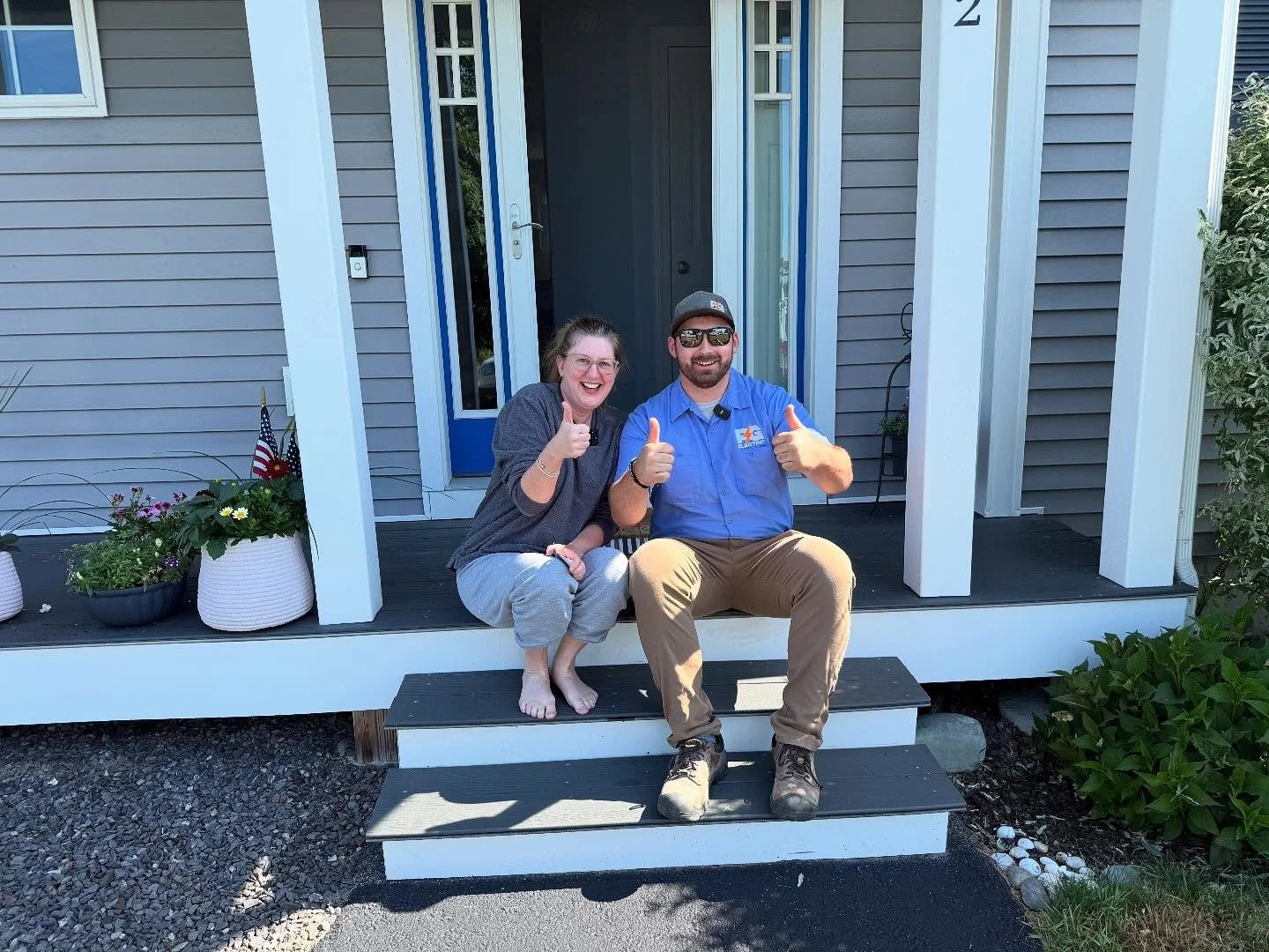 Smiling woman and man sitting on the front steps of a house, giving thumbs up. The woman is barefoot, wearing a gray dress, and the man is in work clothes and boots. The house has gray siding and potted plants near the entrance.