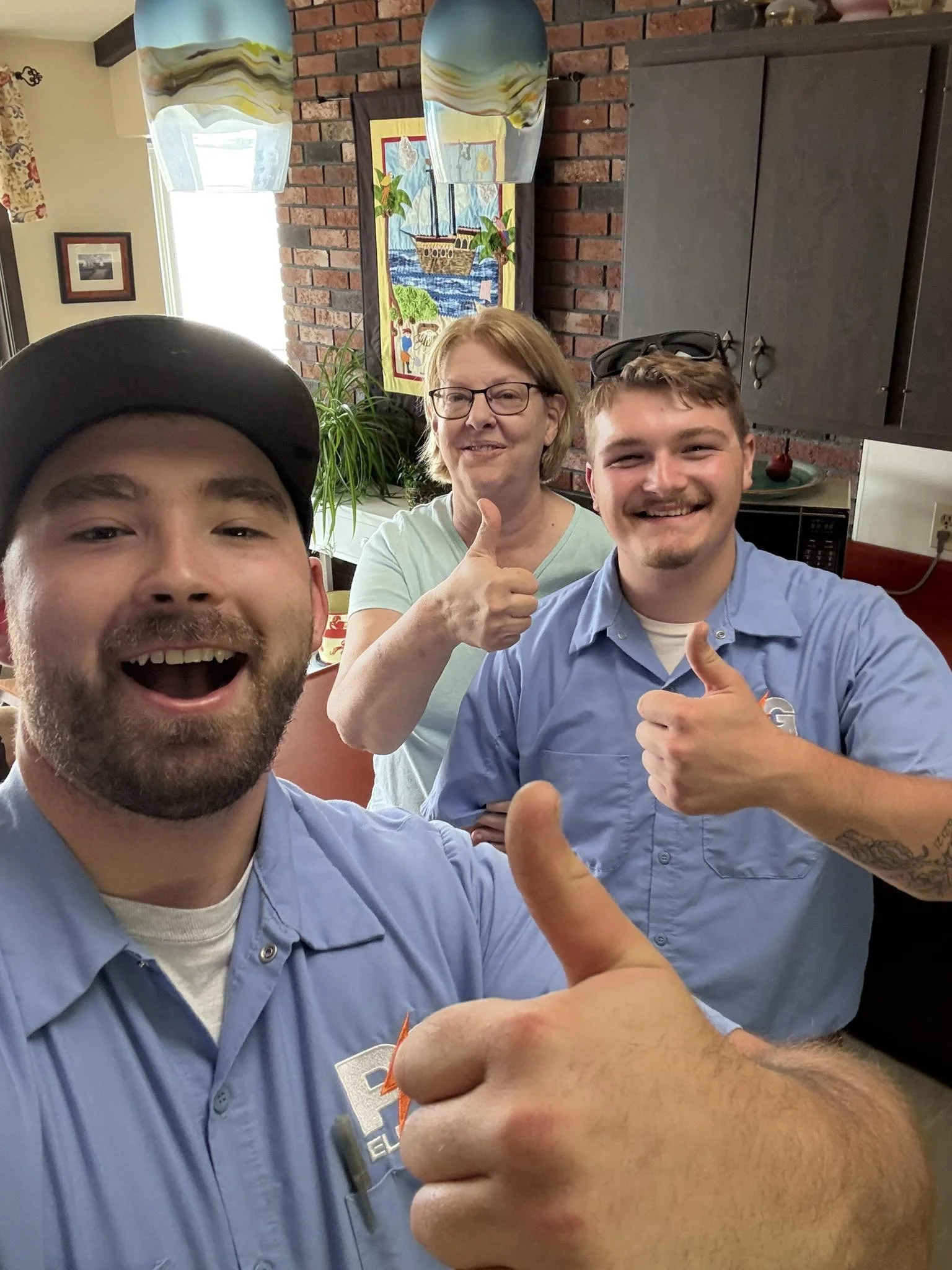 Three people, two young men and a woman, smiling and giving thumbs-up while taking a selfie in a kitchen.
