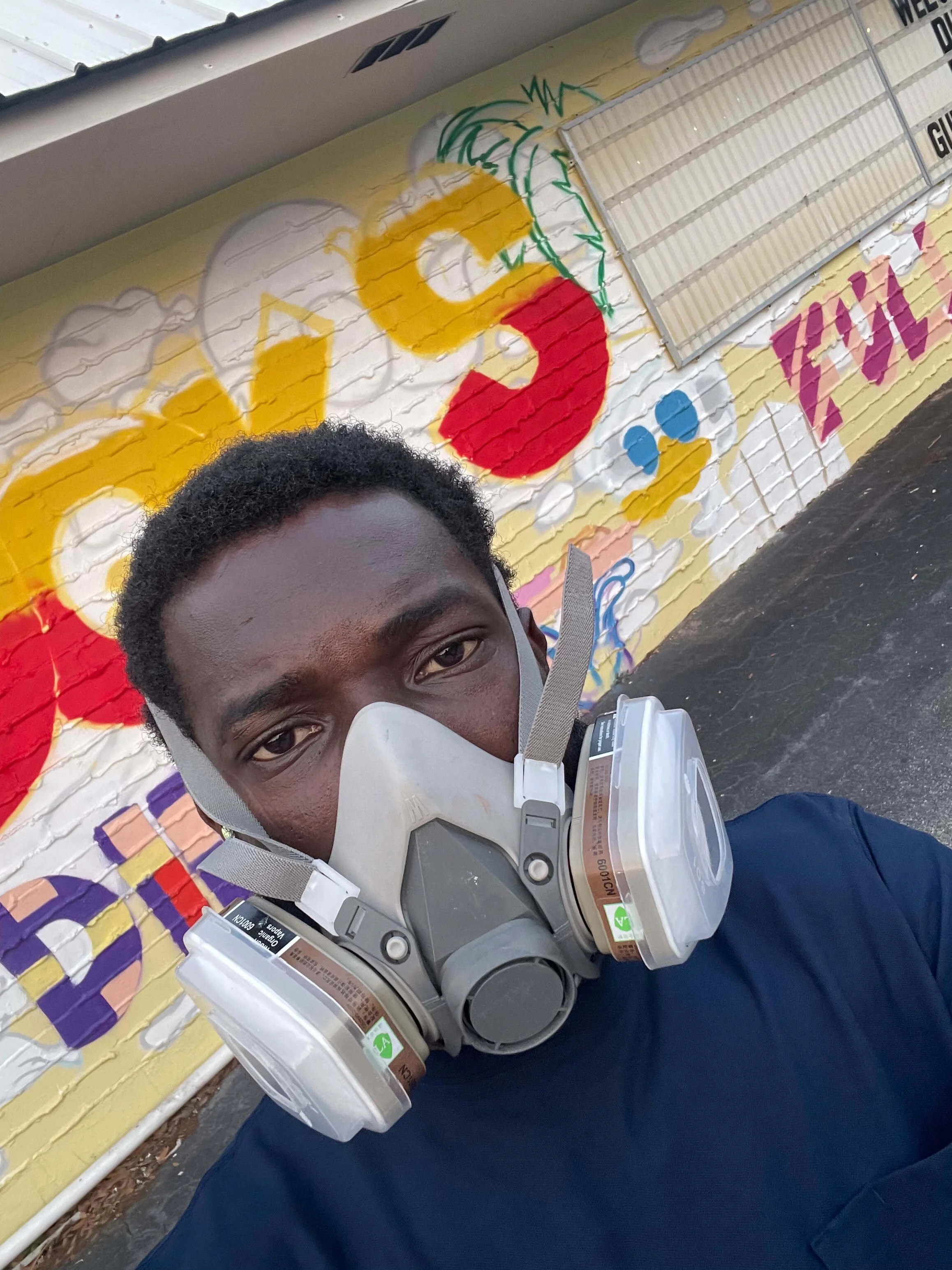 Artist wearing a respirator mask in front of Duck’s Diner mural during painting progress, Gulf Shores AL, 2025.