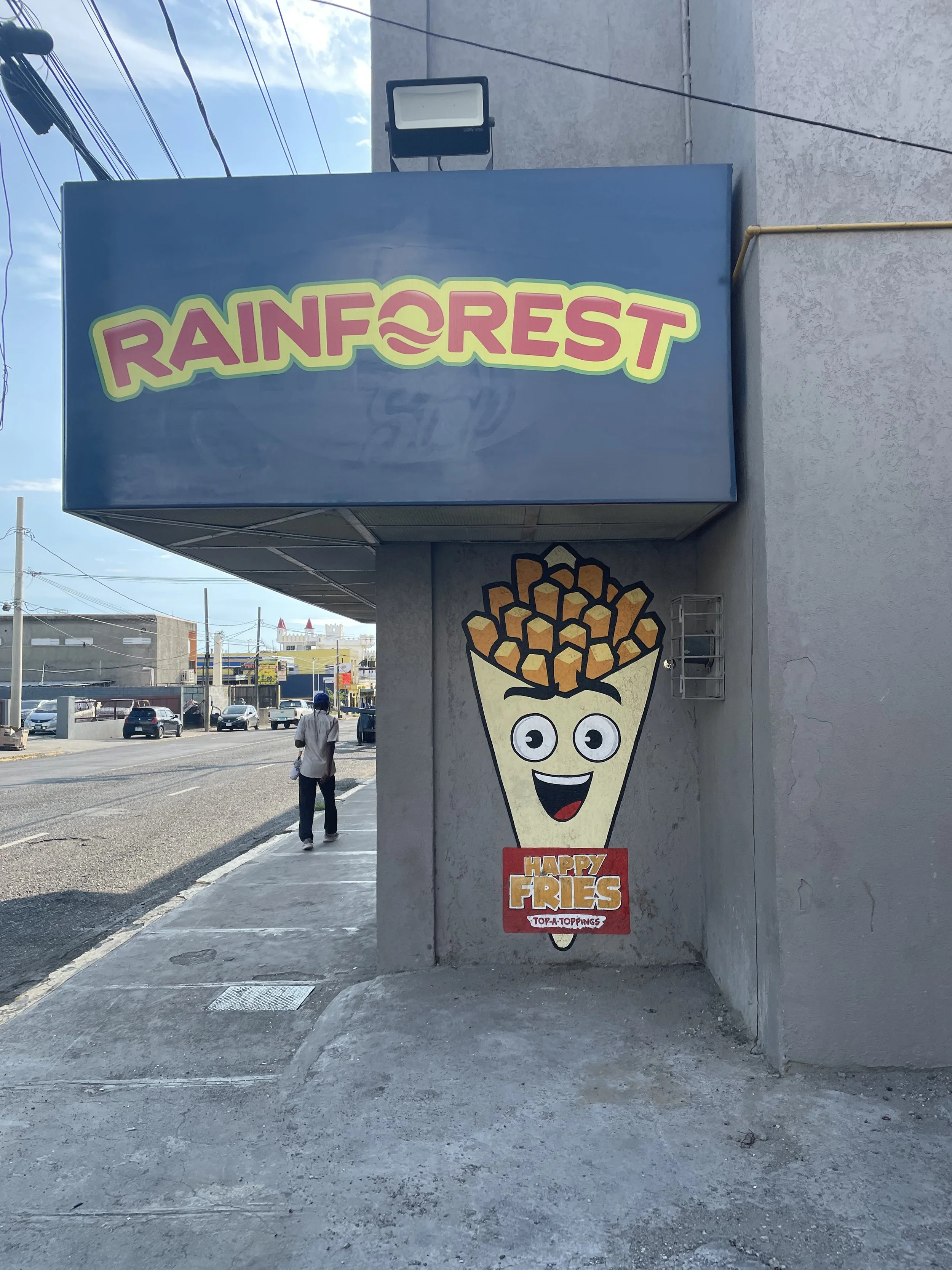 Street view of Rainforest storefront in Kingston, Jamaica, 2025, featuring the Happy Fries mural. The painted mascot shows a cone of fries with a cheerful cartoon face under the Rainforest sign. A lively piece of street branding that highlights Jamai