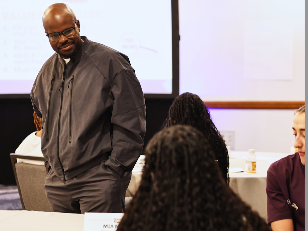A man with glasses wearing a gray jacket standing and engaging with people at a conference or seminar.