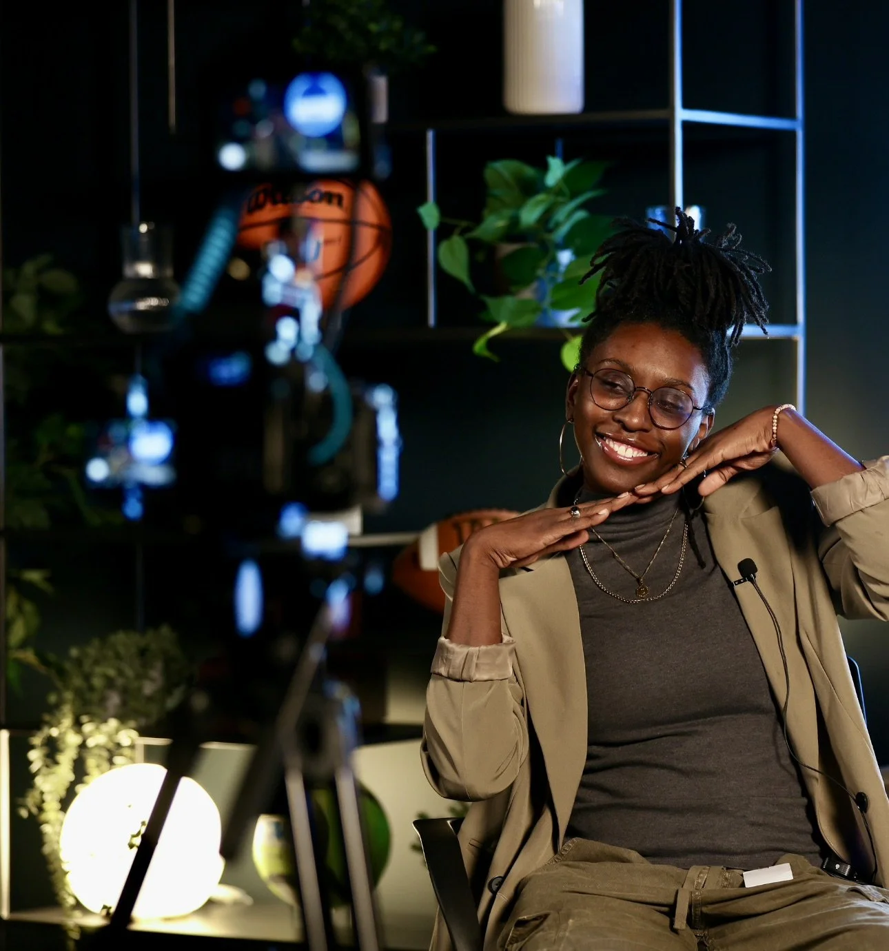 A woman with glasses and a big smile, sitting relaxed in front of a camera, with plants and decorative lights in the background.