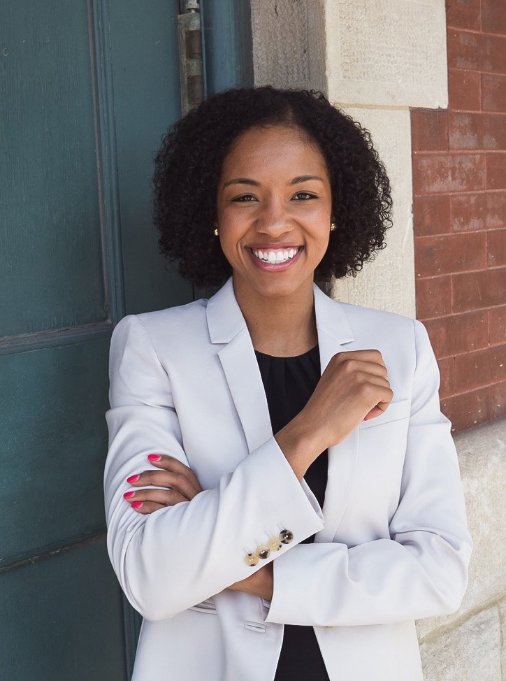 A woman with curly black hair smiling, wearing a white blazer over a black top, standing outdoors against a brick and white wall.
